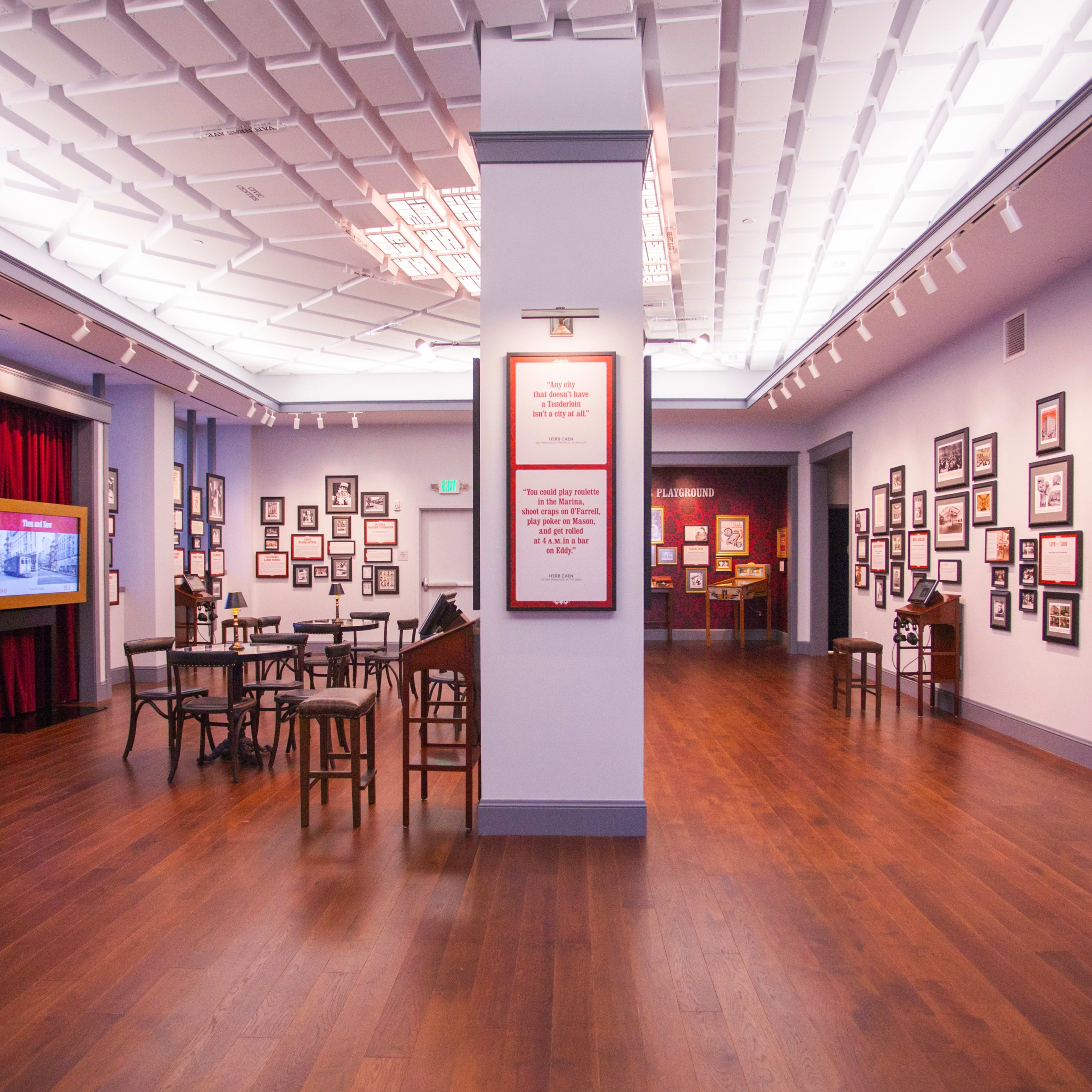 An interior of a museum or gallery with wooden flooring, framed pictures on the walls, and several tables arranged in the space.