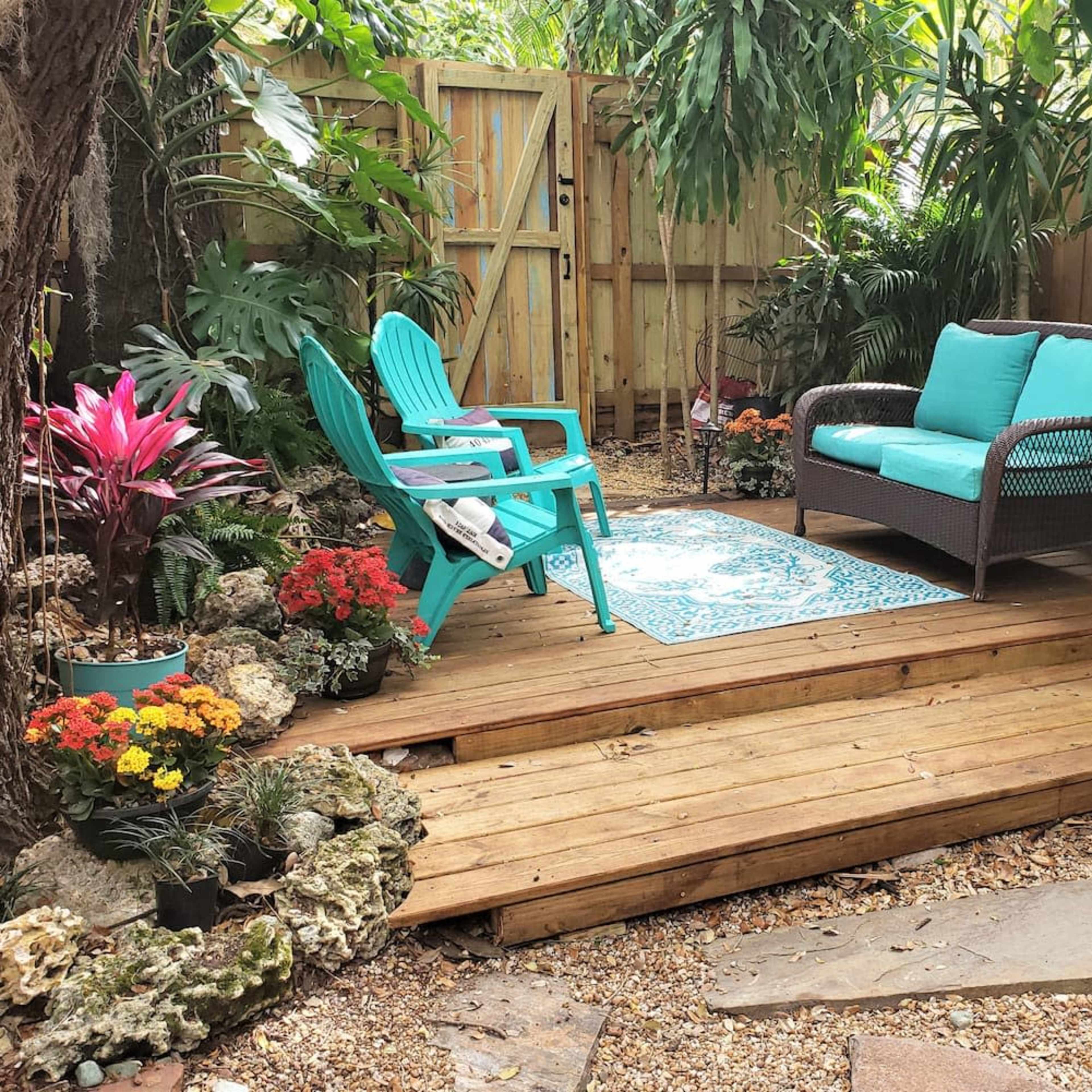 A wooden deck in a garden area, featuring a turquoise sofa and an Adirondack chair surrounded by potted plants and flowers.