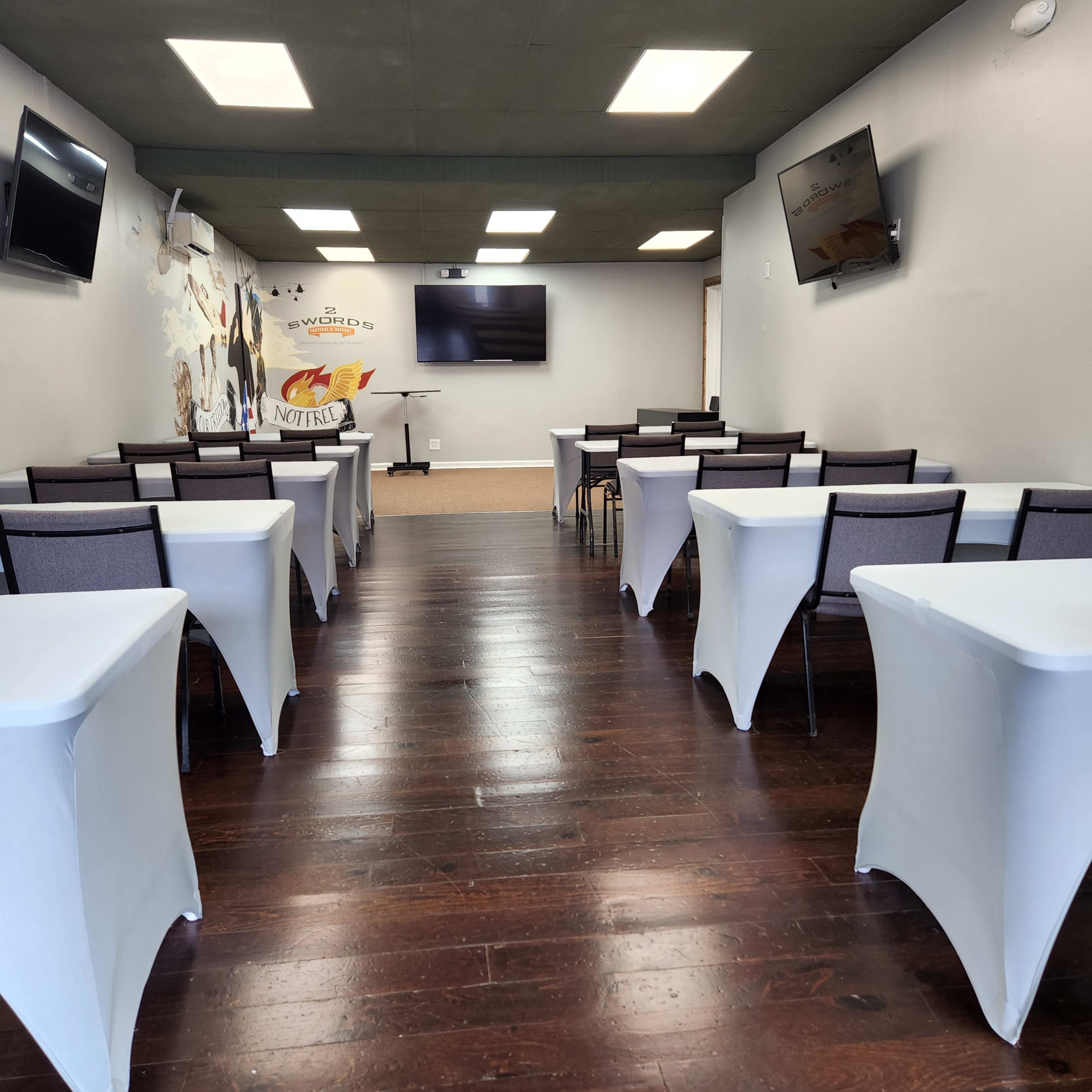 A room set up with multiple tables covered in white linens and equipped with chairs, along with several wall-mounted televisions.