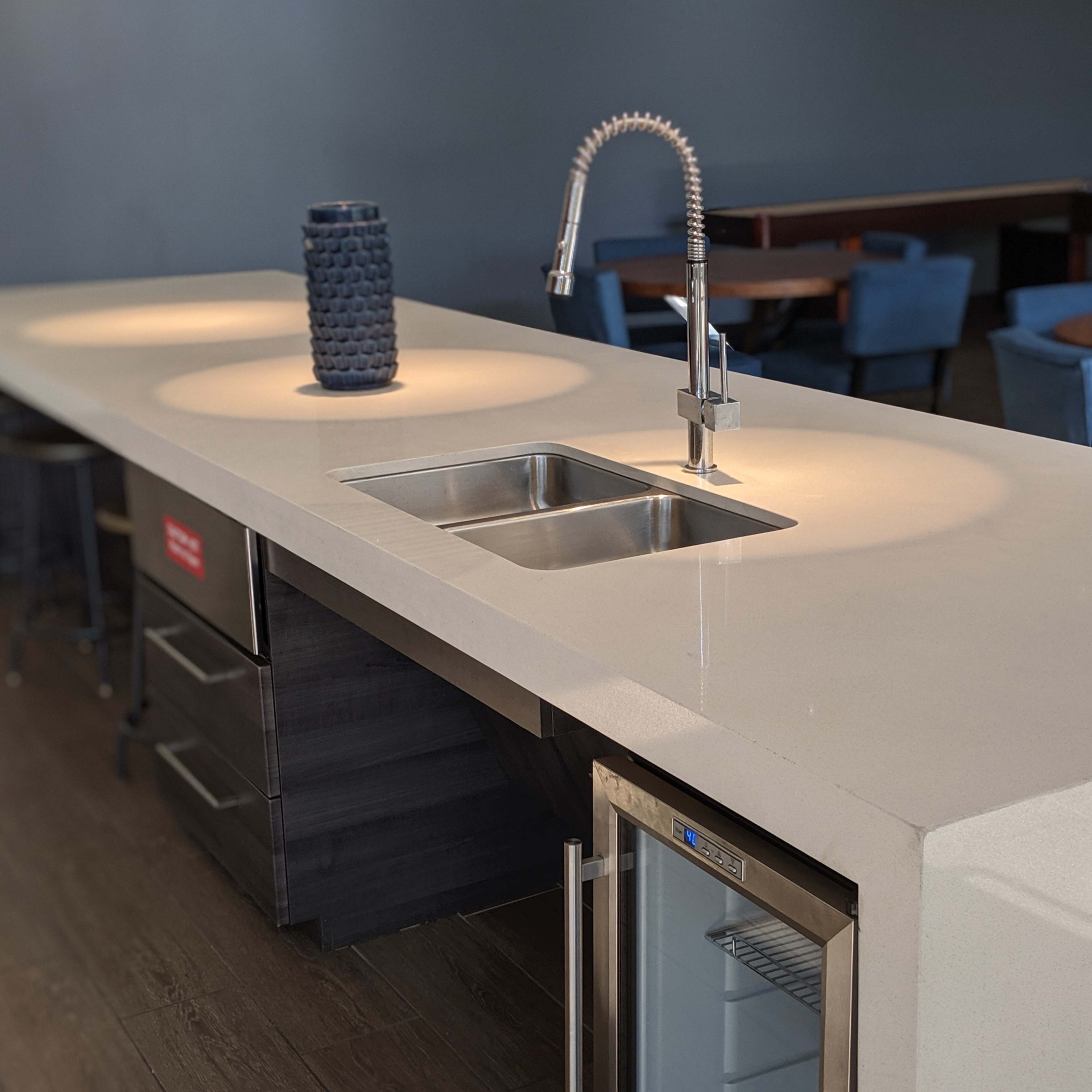 A modern kitchen countertop featuring a stainless steel sink, a faucet, and a small refrigerator, with ambient lighting highlighting the space.