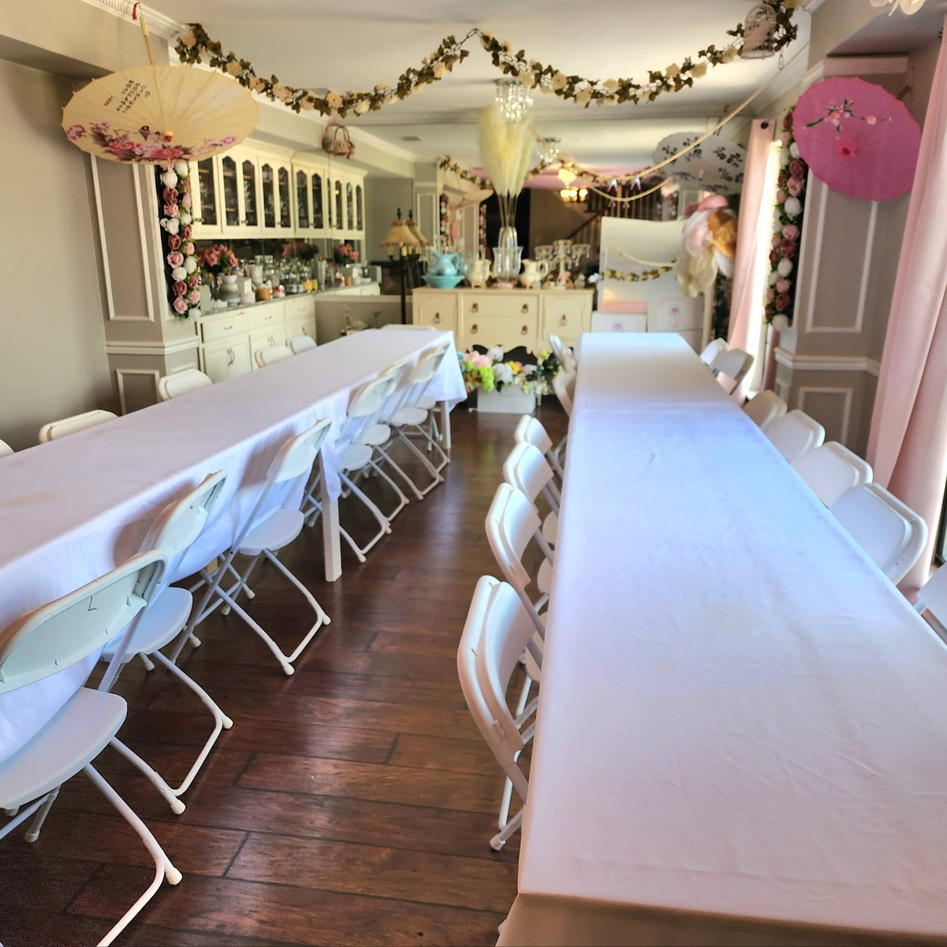 A decorated room with two long tables covered in white tablecloths, arranged with folding chairs along the sides, and floral decorations hanging overhead.