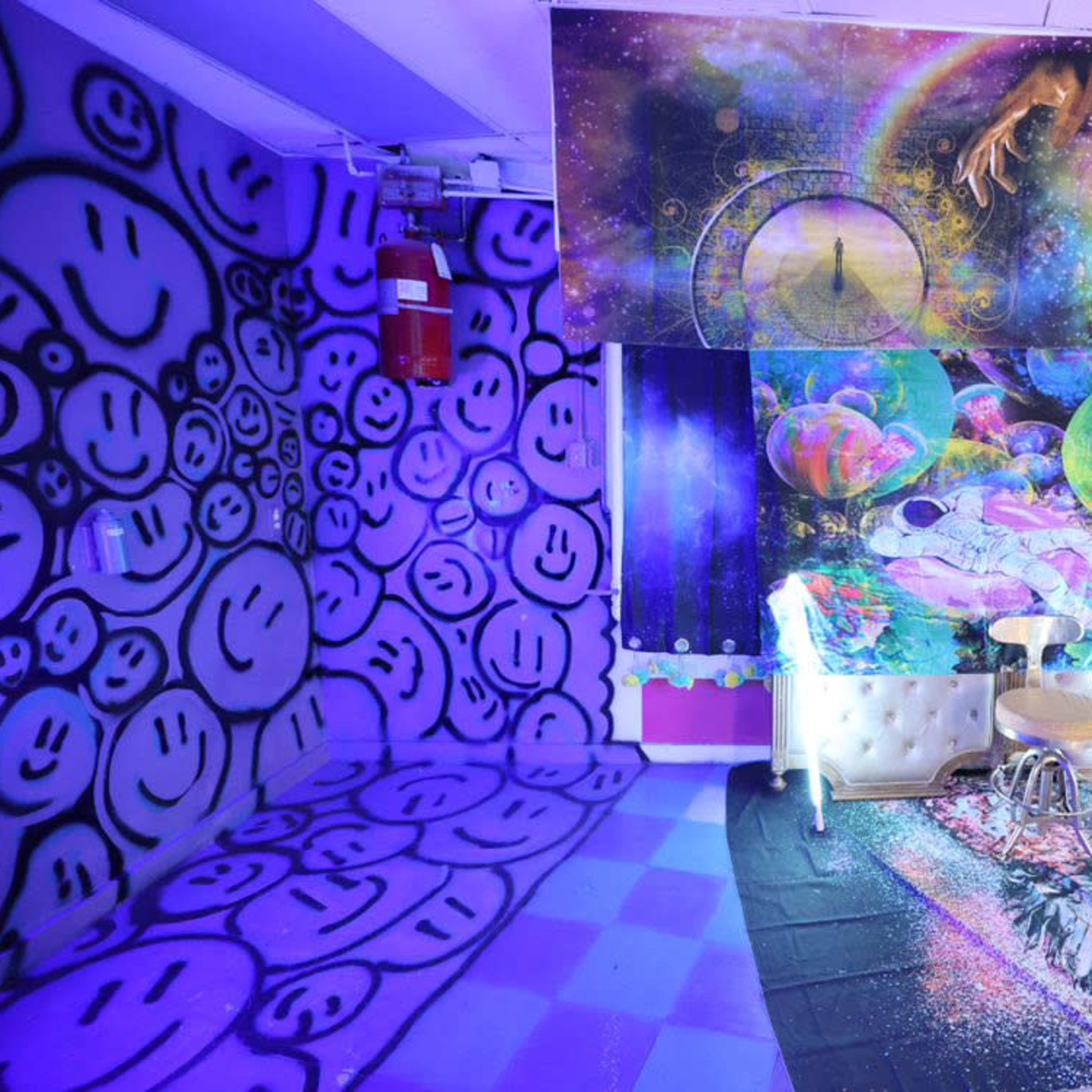 A room decorated with vibrant graffiti of smiley faces on one wall, colorful cosmic-themed art on the ceiling, and a white couch with a stool positioned in front of it.