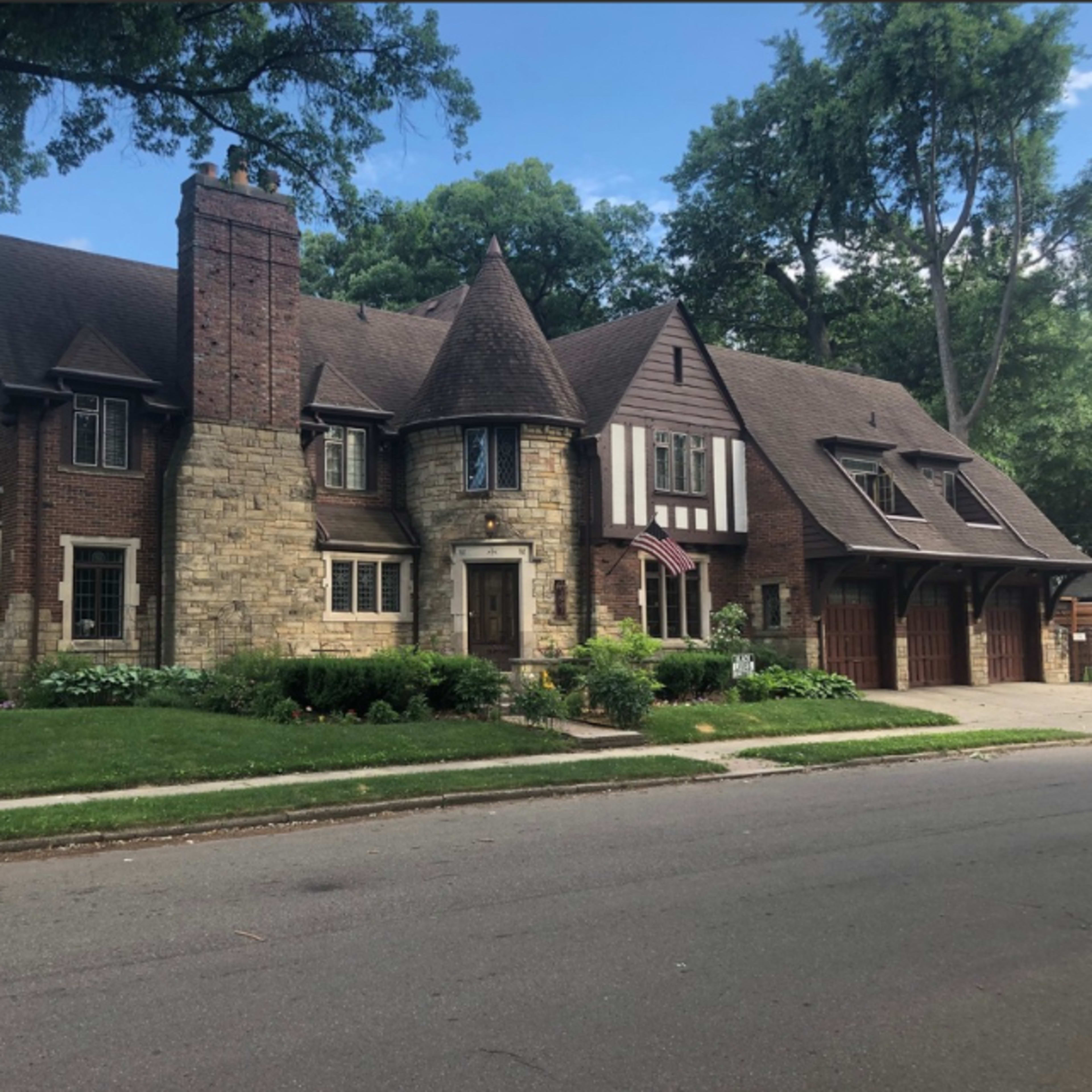 A large, two-story, Tudor-style house with a combination of brick and stone exteriors, featuring a prominent chimney and multiple gabled roofs.