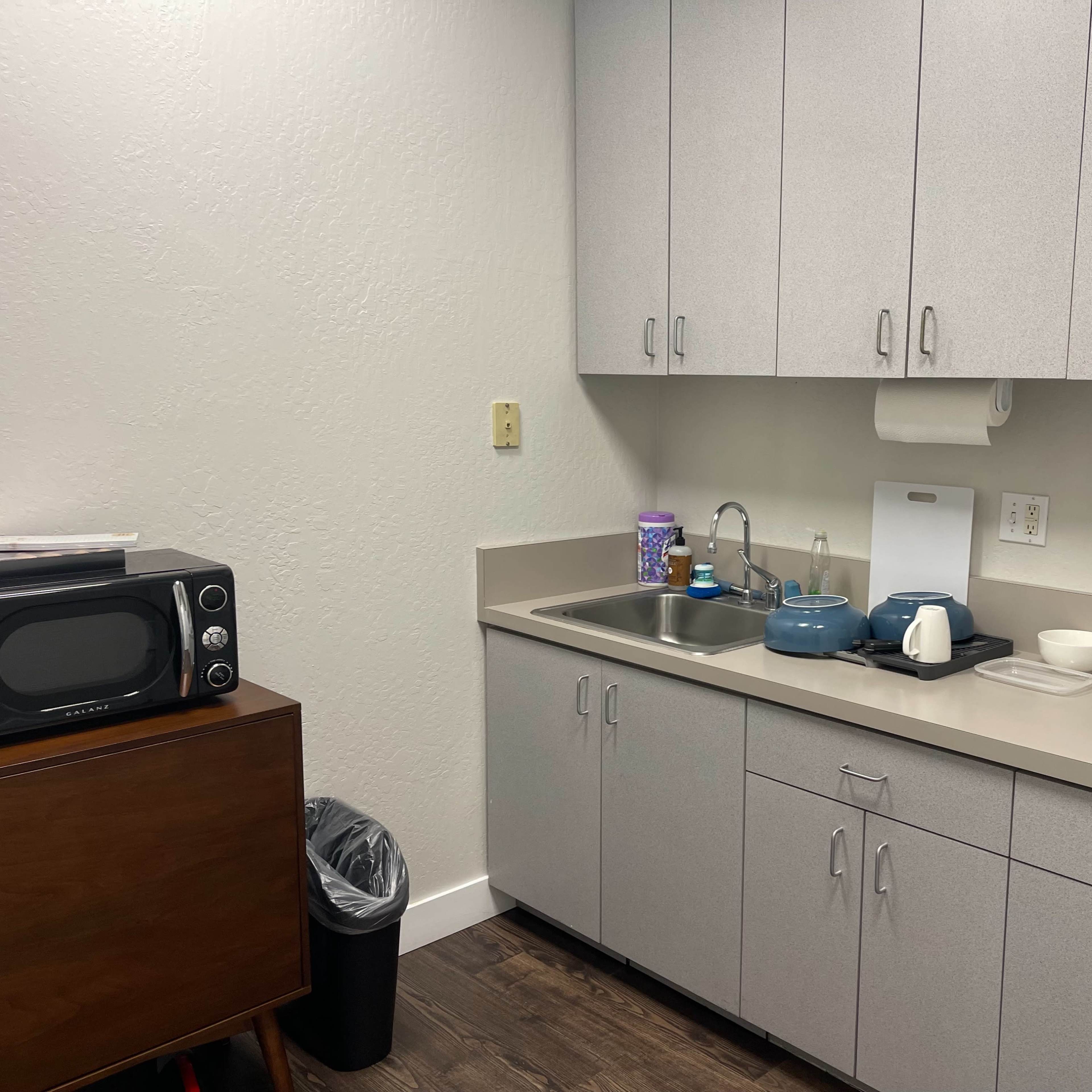 A small kitchen area with a microwave, a sink, blue cookware, and a trash can in the corner.