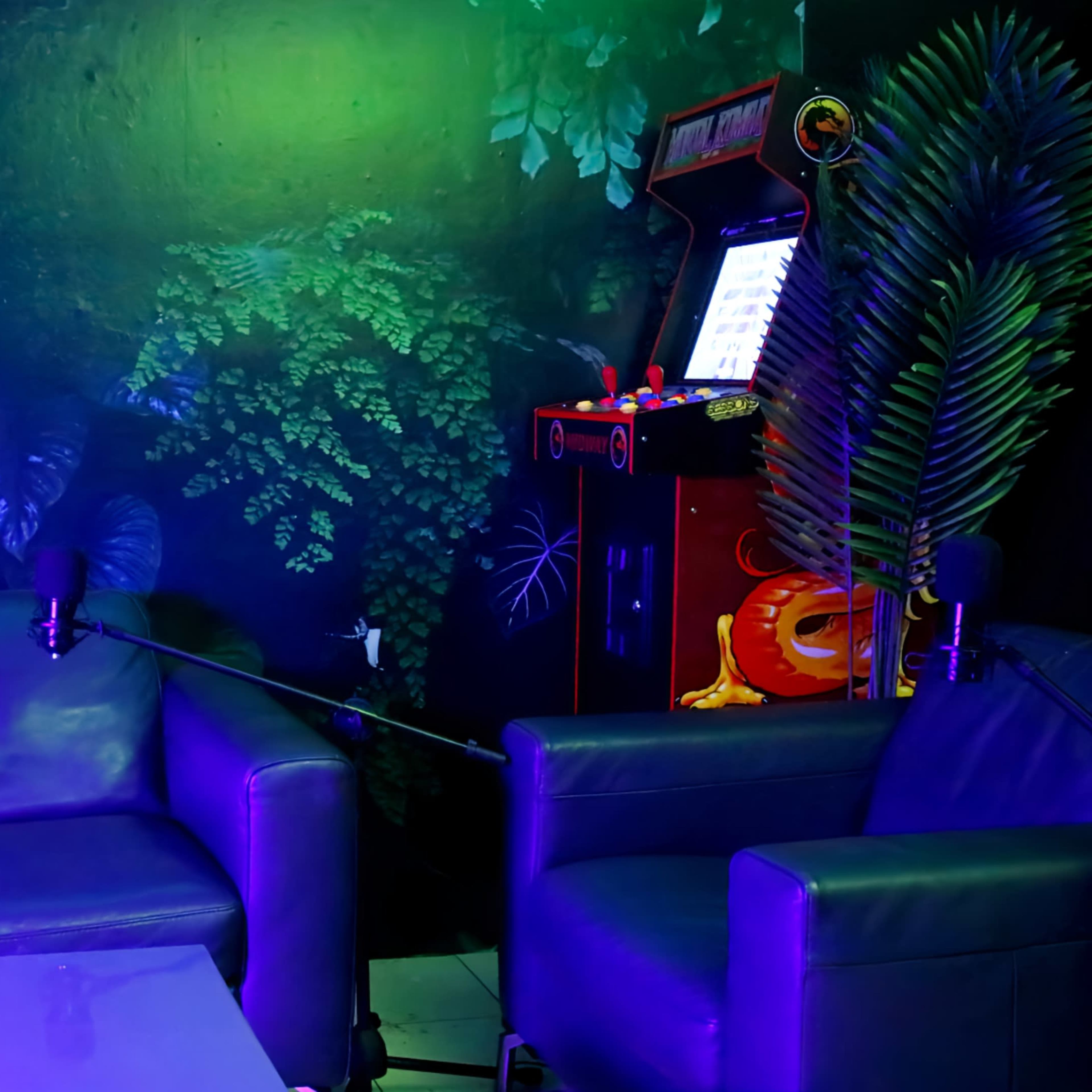 The scene features a dimly lit room with a dark green plant-themed mural, a blue leather couch, a white table, and a vintage arcade machine in the corner.