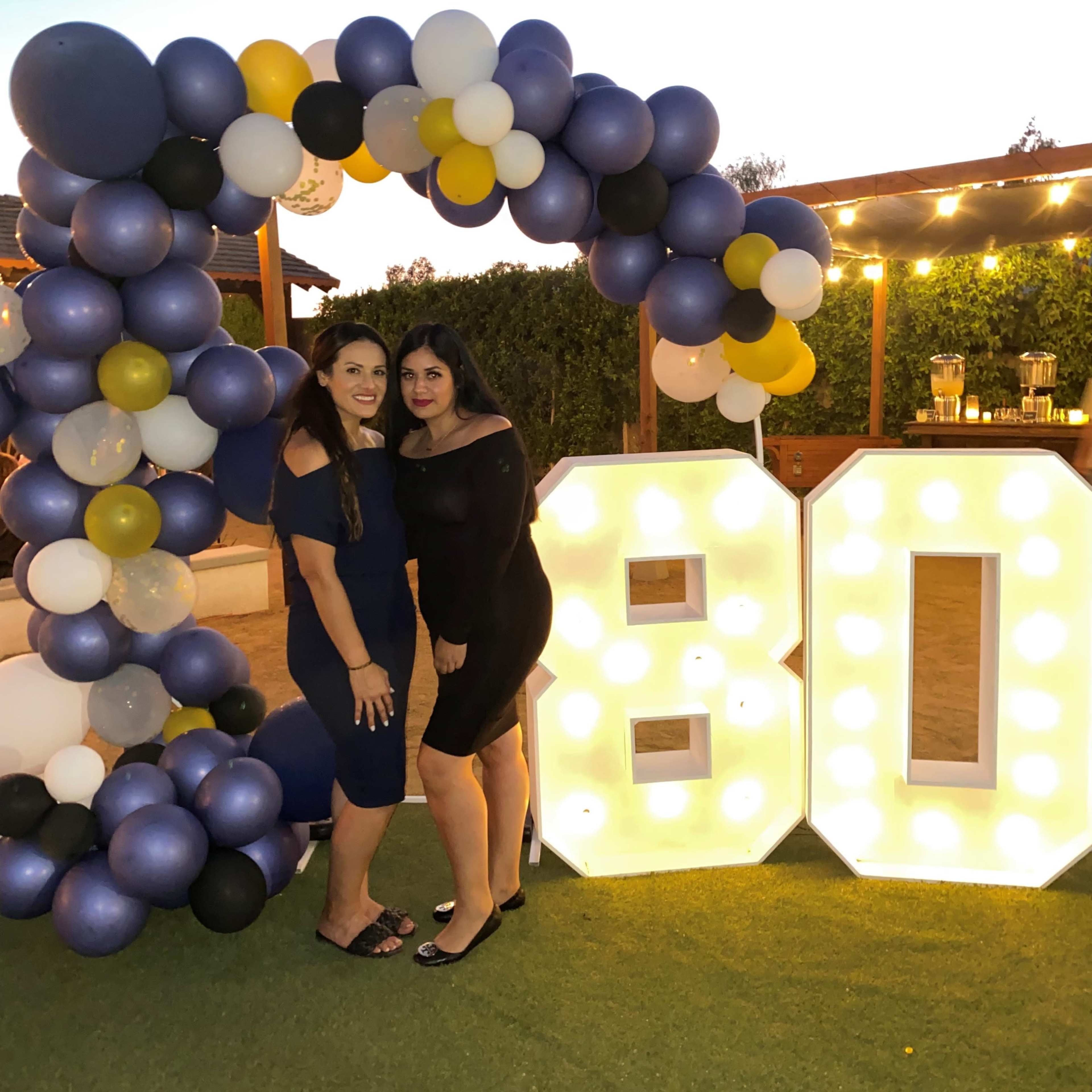 Two women pose together in front of large illuminated "80" numbers, surrounded by a decorative arch of balloons.