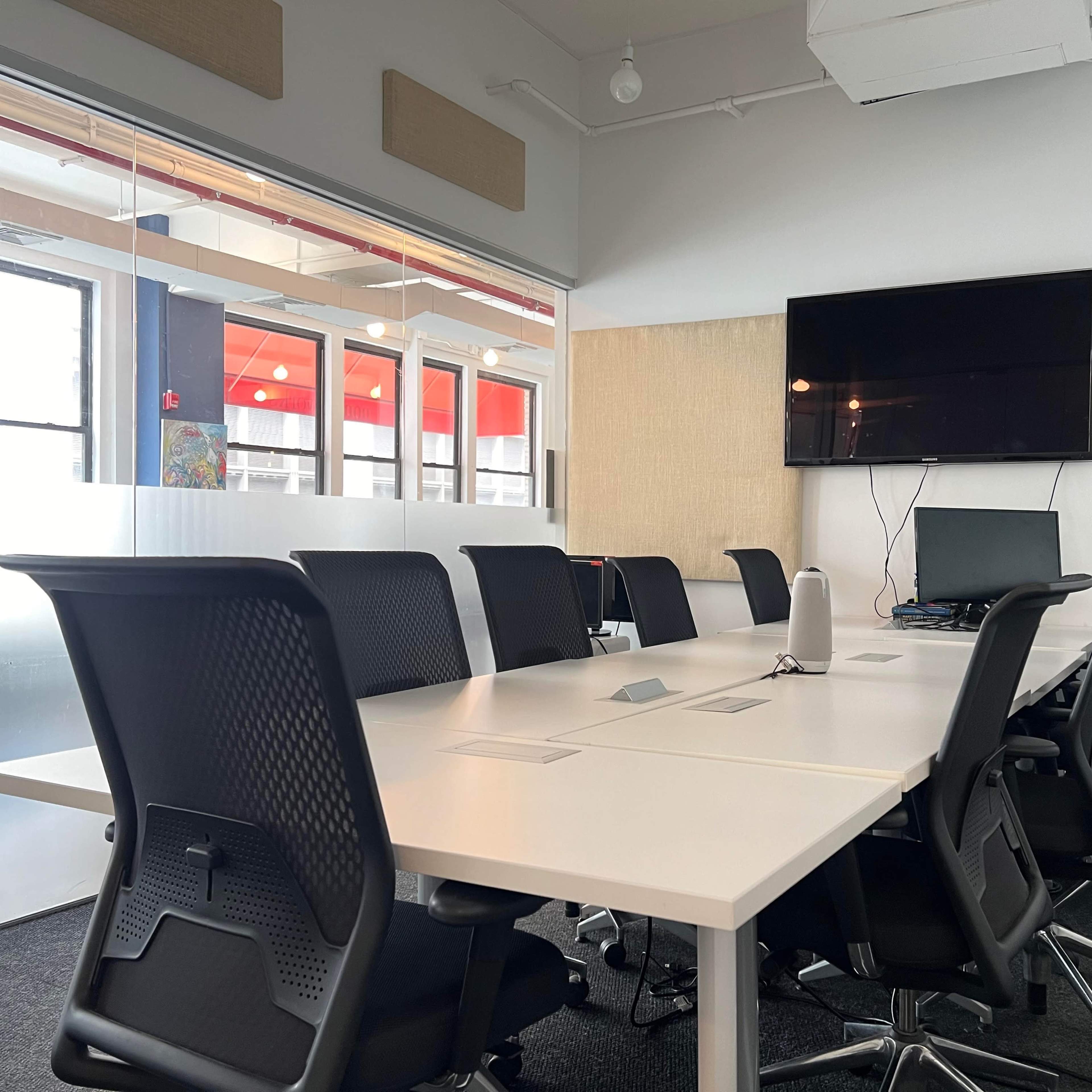 A modern conference room features a large white table surrounded by black office chairs, with a wall-mounted TV and computer setup in the background.