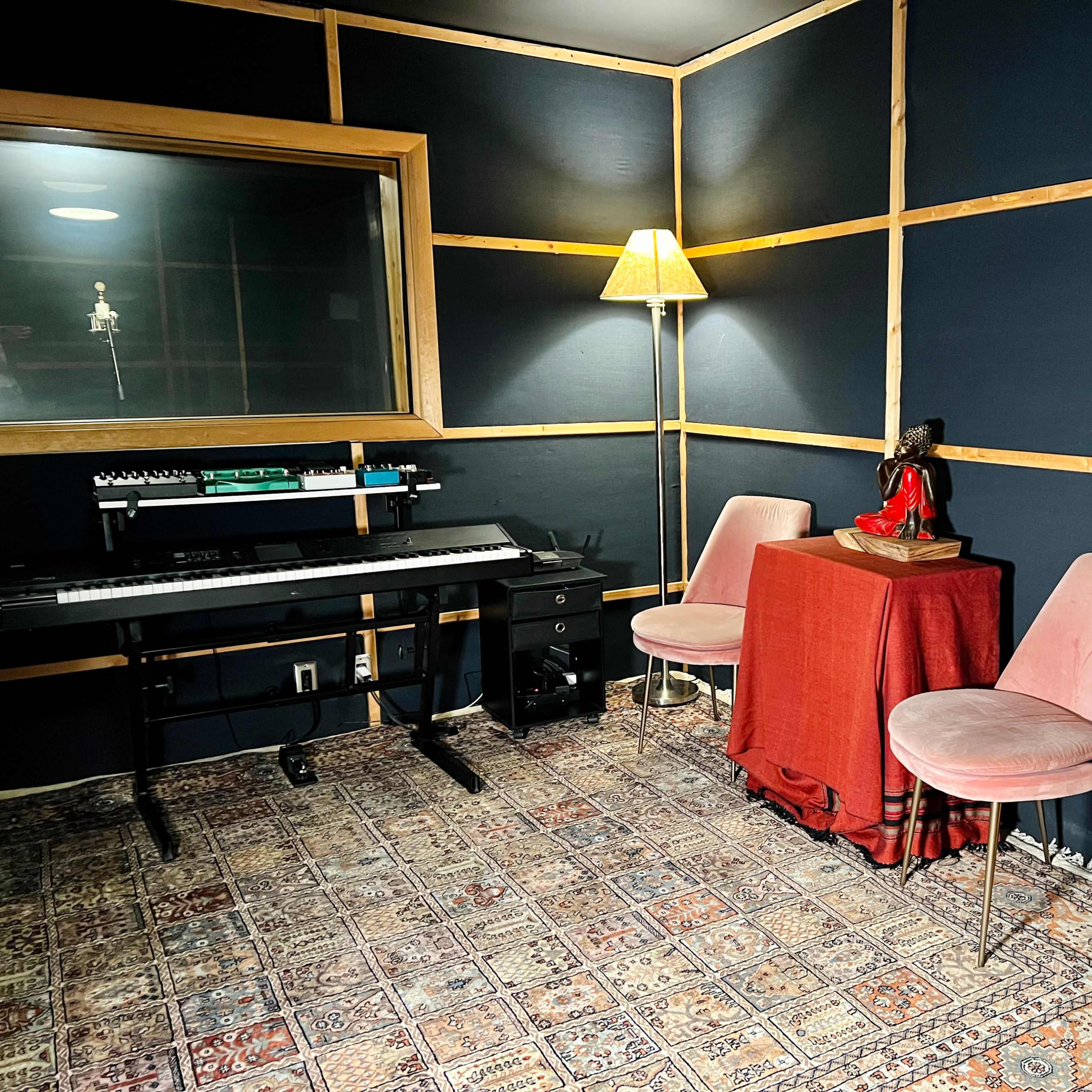 A small music studio featuring a keyboard on a stand, two pink chairs, a red table, a lamp, a statue, and an acoustic guitar leaning against the wall.