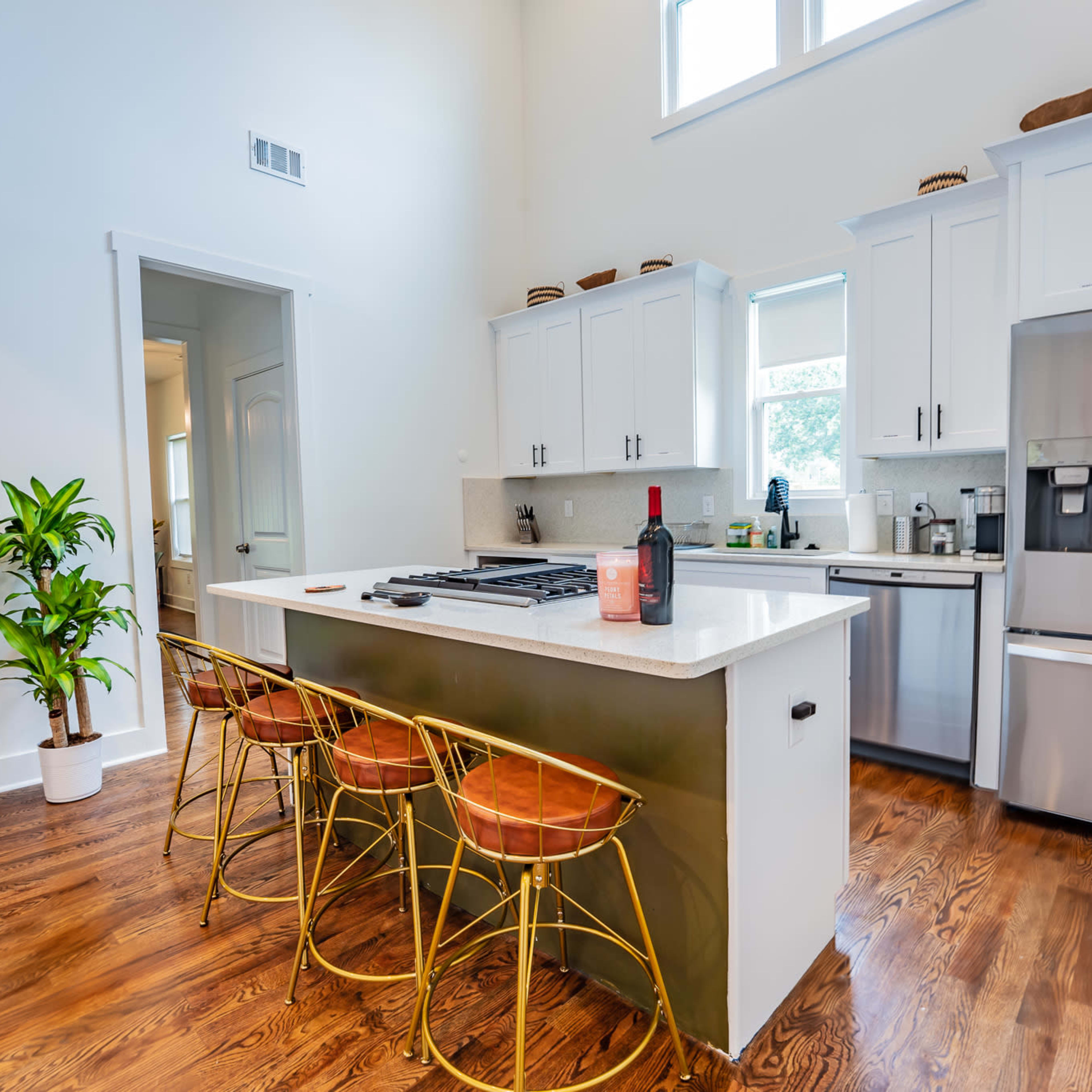 A modern kitchen features a central island with three gold bar stools, stainless steel appliances, and a large window providing natural light.