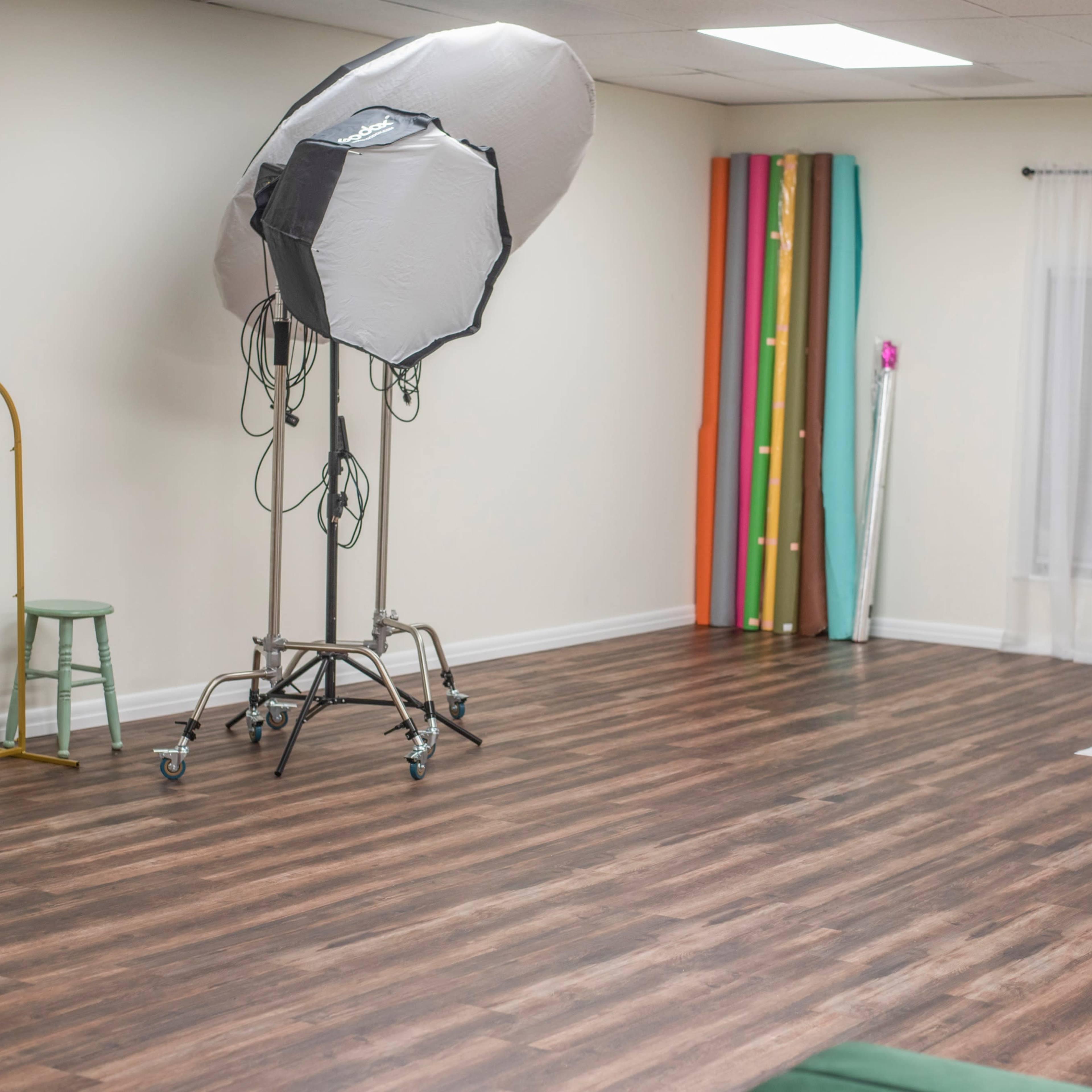 A photography studio with a large light umbrella, a stool, a full-length mirror, and colorful backdrop rolls along the wall.
