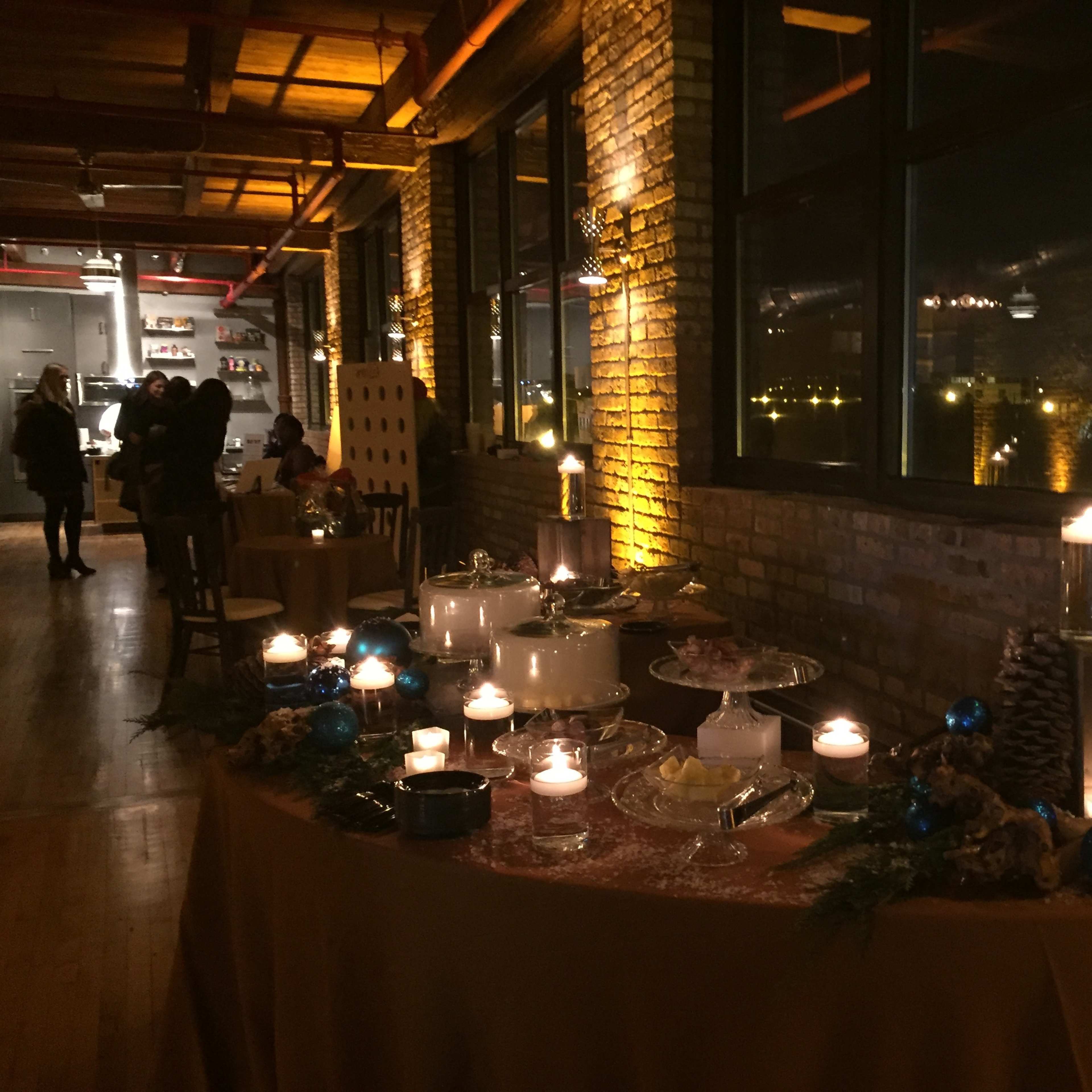A table adorned with various desserts and candles is set against a backdrop of a warmly lit, industrial-style venue with large windows.