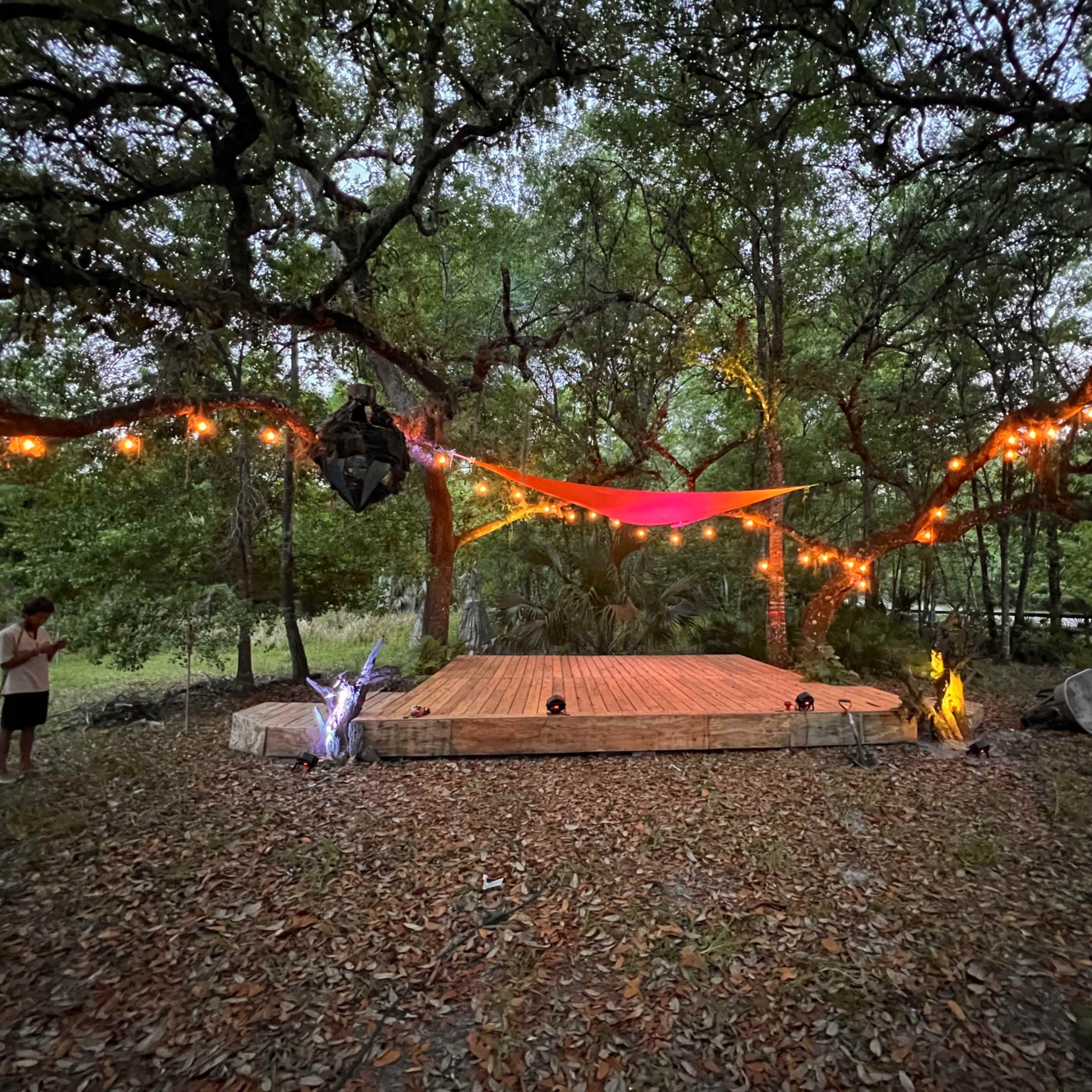 A wooden stage is set under trees adorned with string lights and colorful fabric, surrounded by a natural forest backdrop.