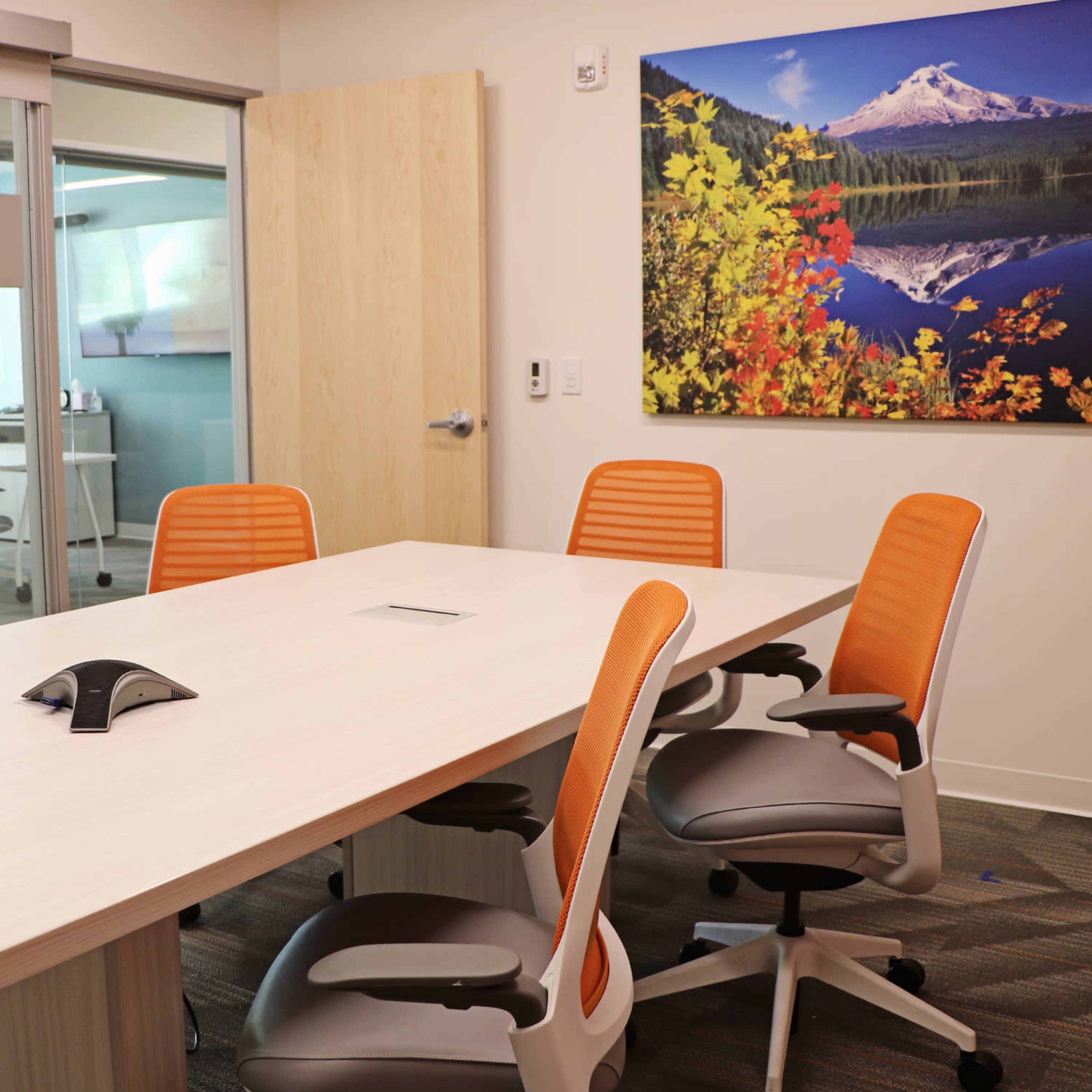 A modern conference room with a rectangular table, four orange office chairs, and a large wall-mounted nature landscape artwork.