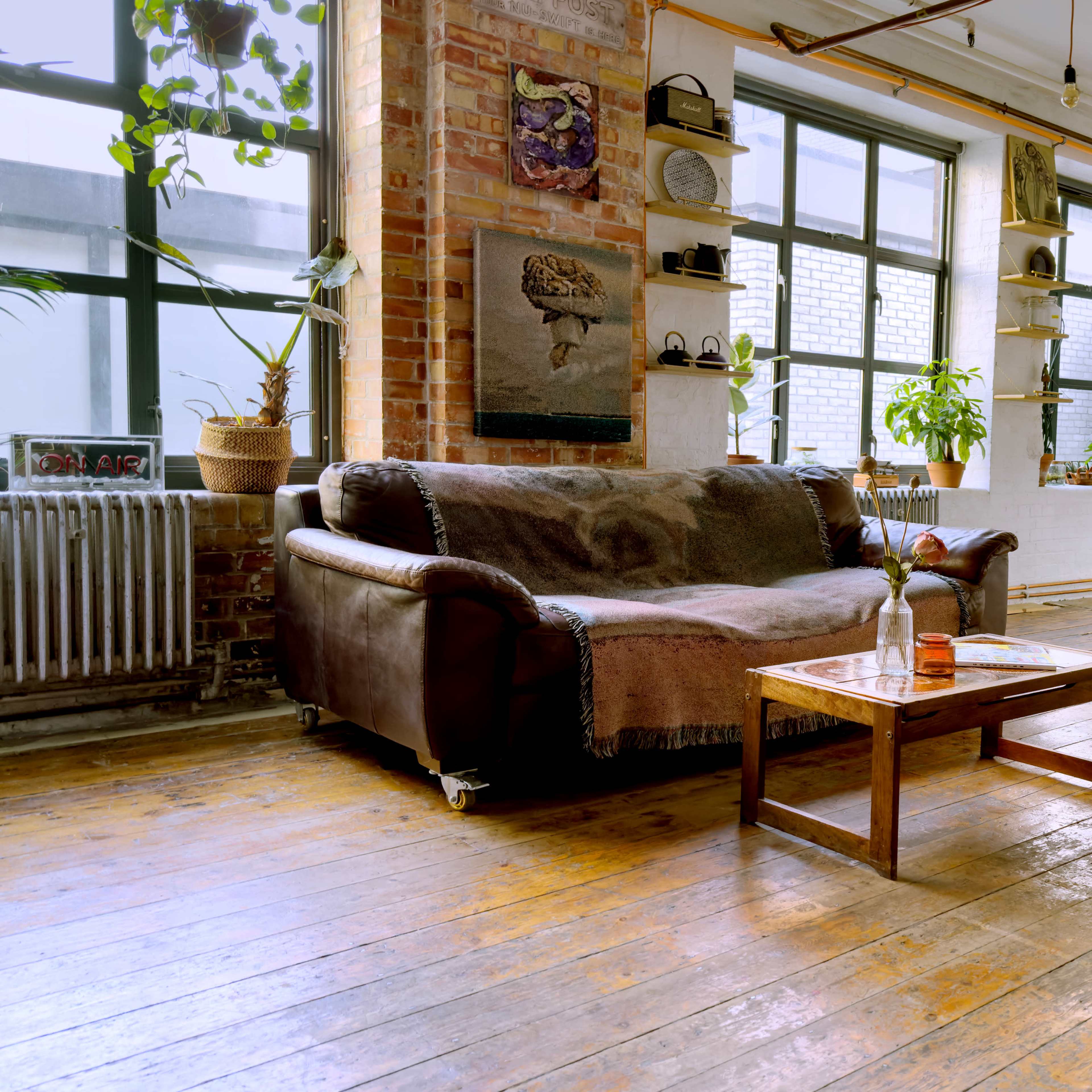 A spacious, sunlit room featuring a brown leather sofa, a wooden coffee table, and large windows with plants and decorative items placed throughout.