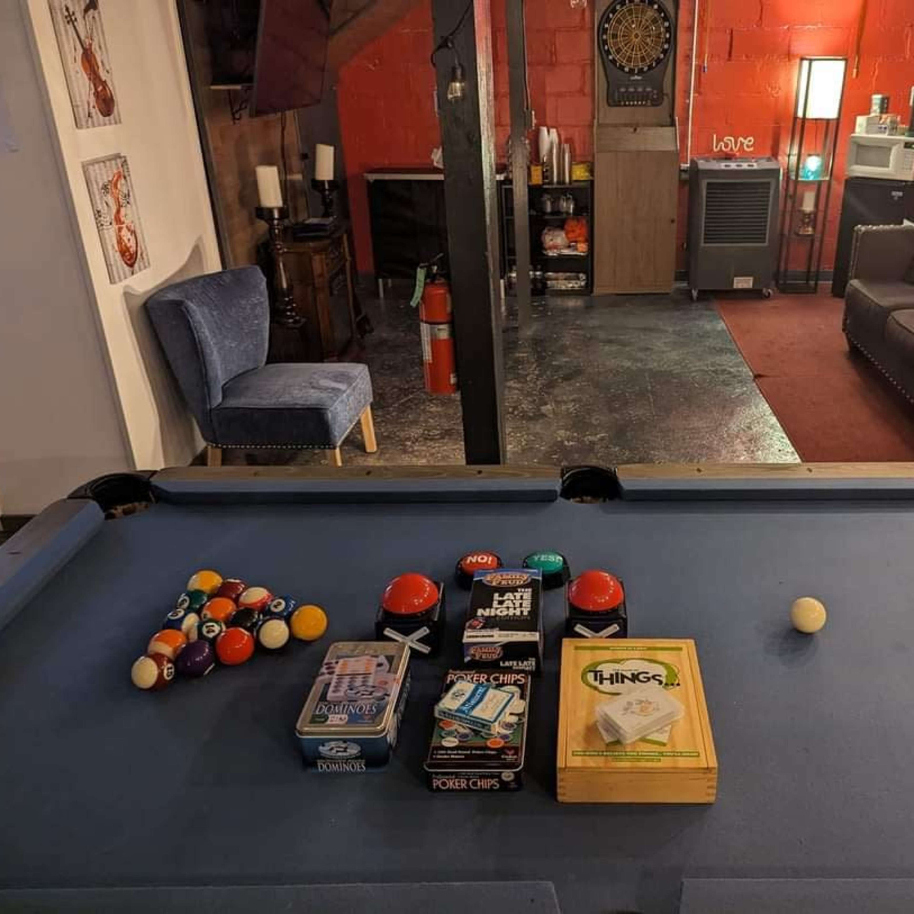 A pool table is set up in a room with red walls, featuring games and items related to billiards and board games, with a couch and dartboard visible in the background.