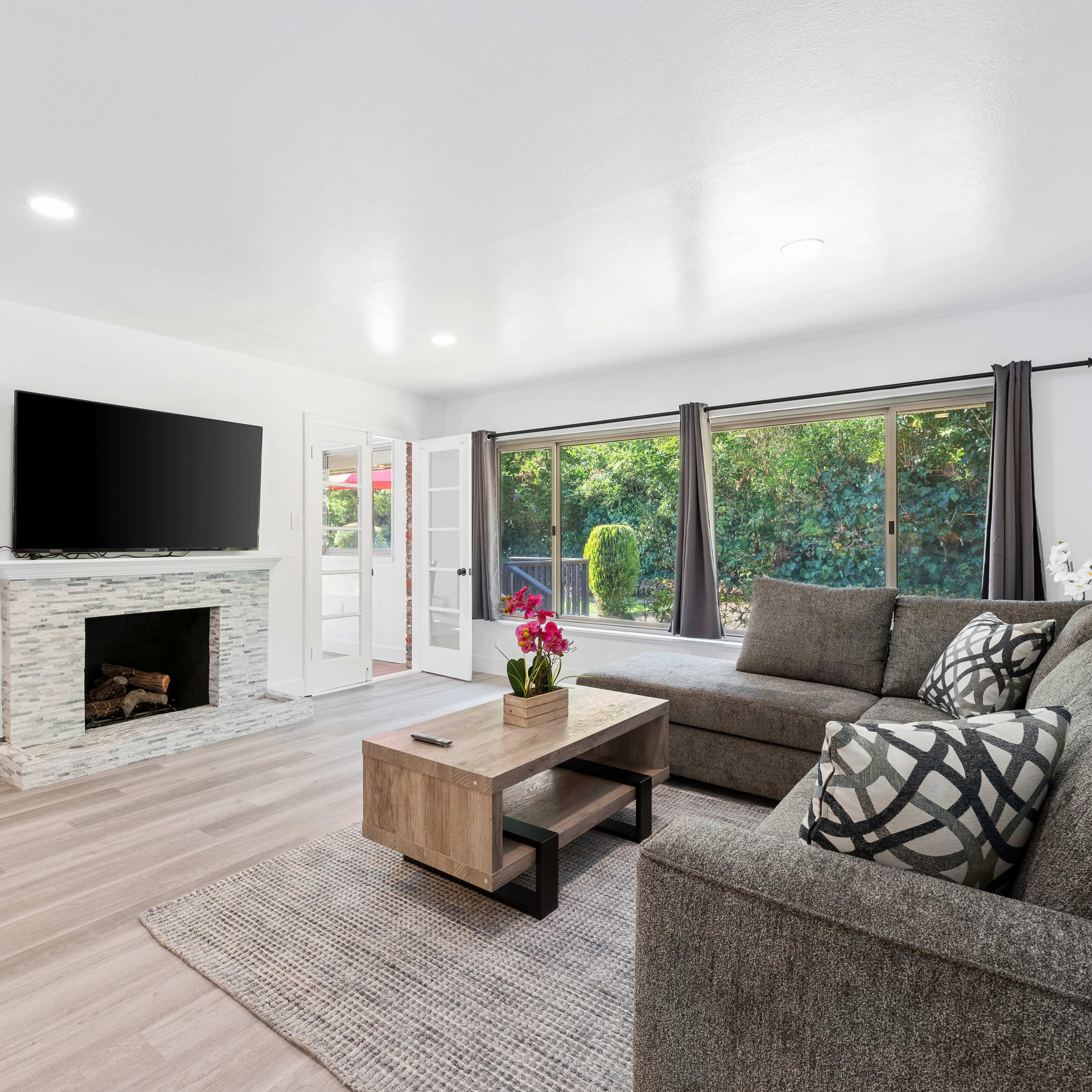 A modern living room features a gray sofa, a wooden coffee table, a stone fireplace, and large windows overlooking a garden.