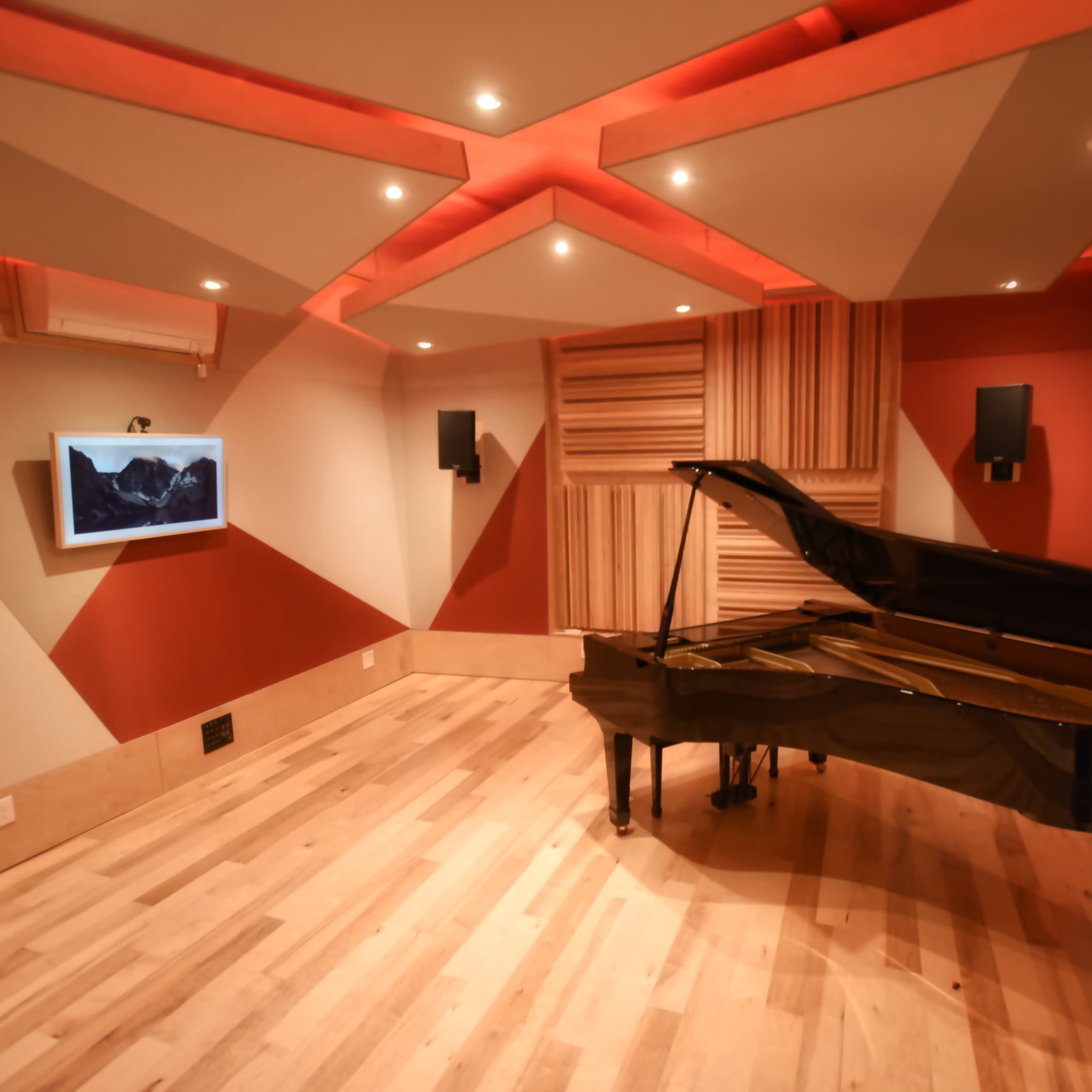 A black grand piano is positioned in a soundproof room with patterned red and beige walls, wooden flooring, and ambient lighting.