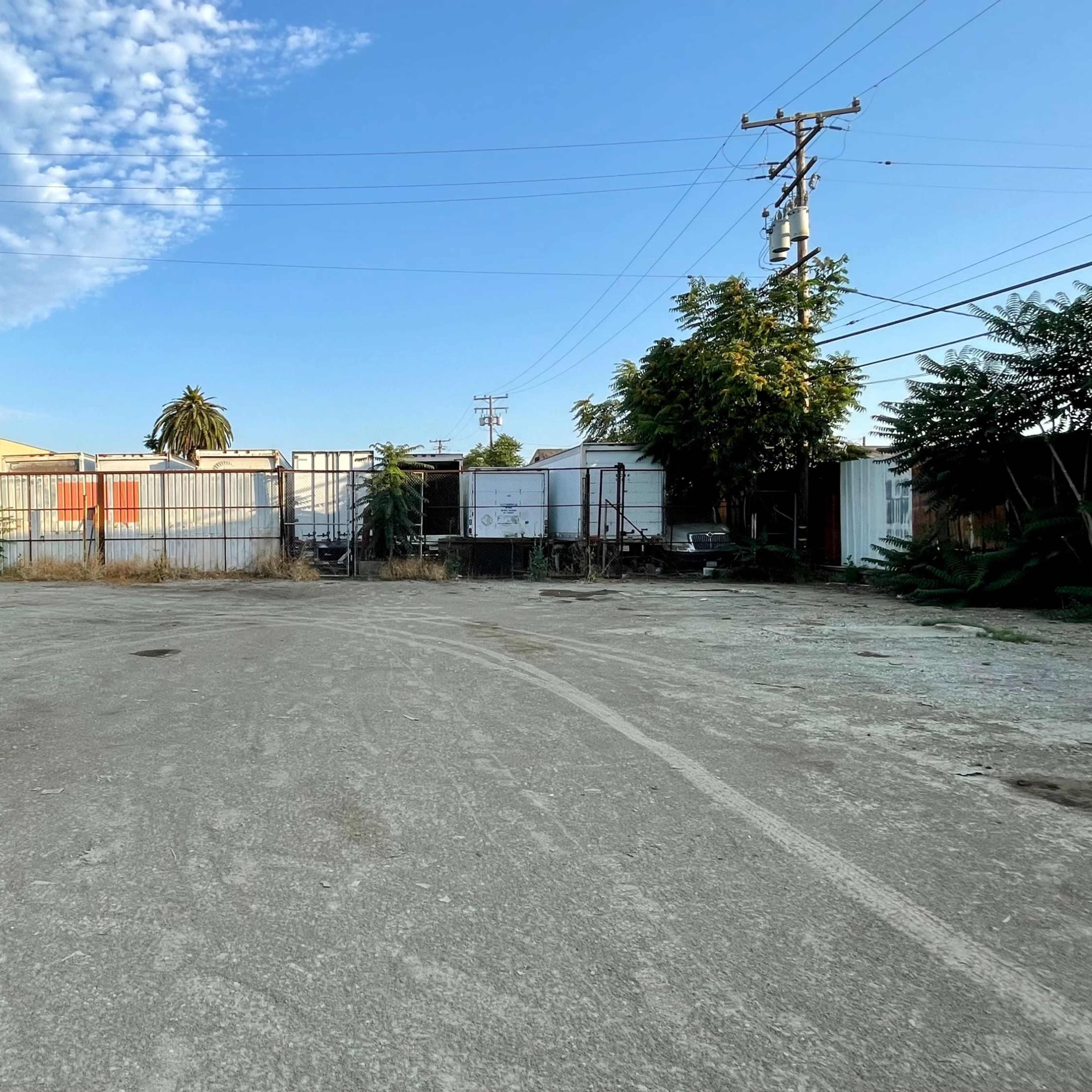 A vacant lot with gravel and sparse vegetation, enclosed by industrial fencing and power lines against a clear blue sky.