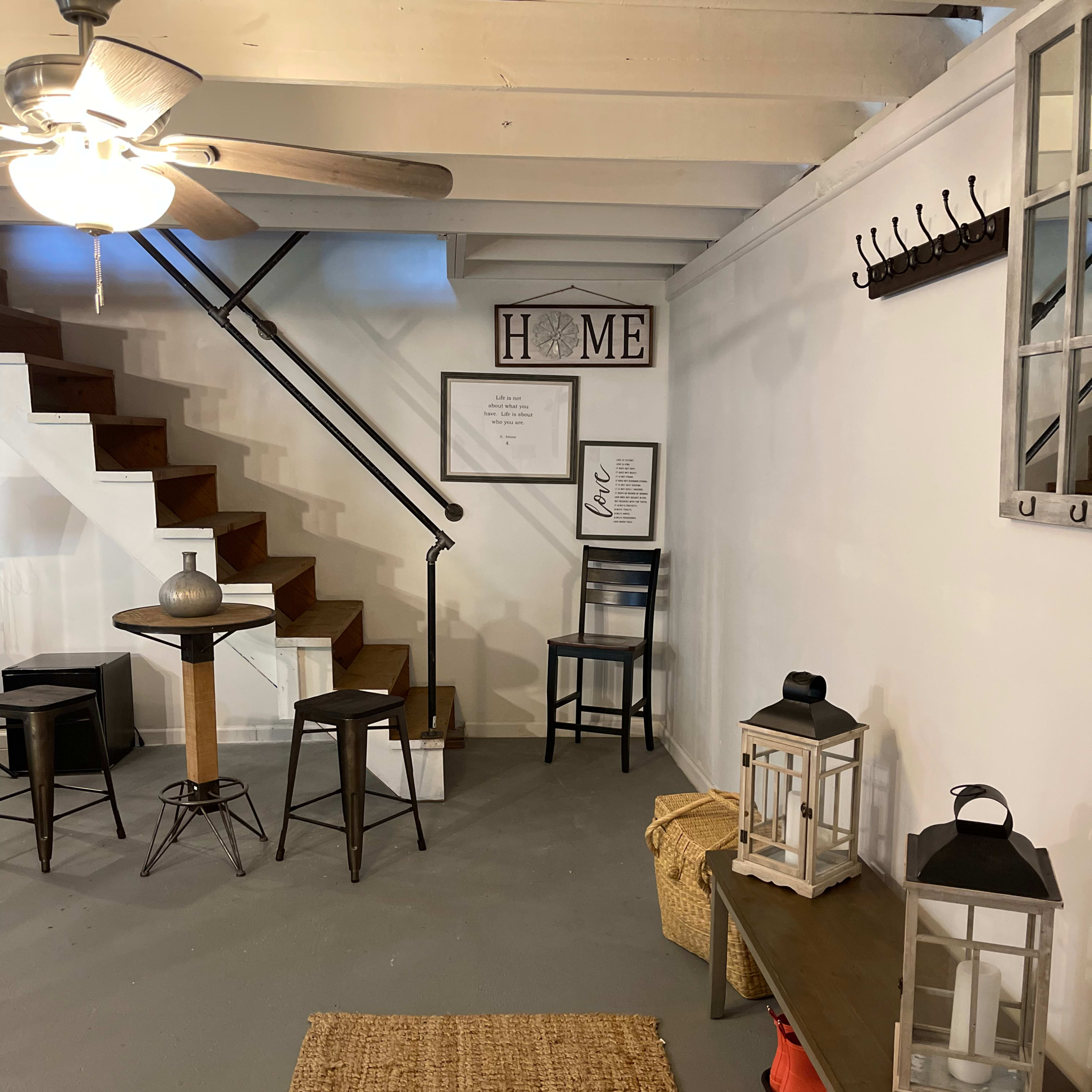 A basement area with a staircase, a small table and chairs, wall decor featuring the word "HOME," and a couple of lanterns on a console table.