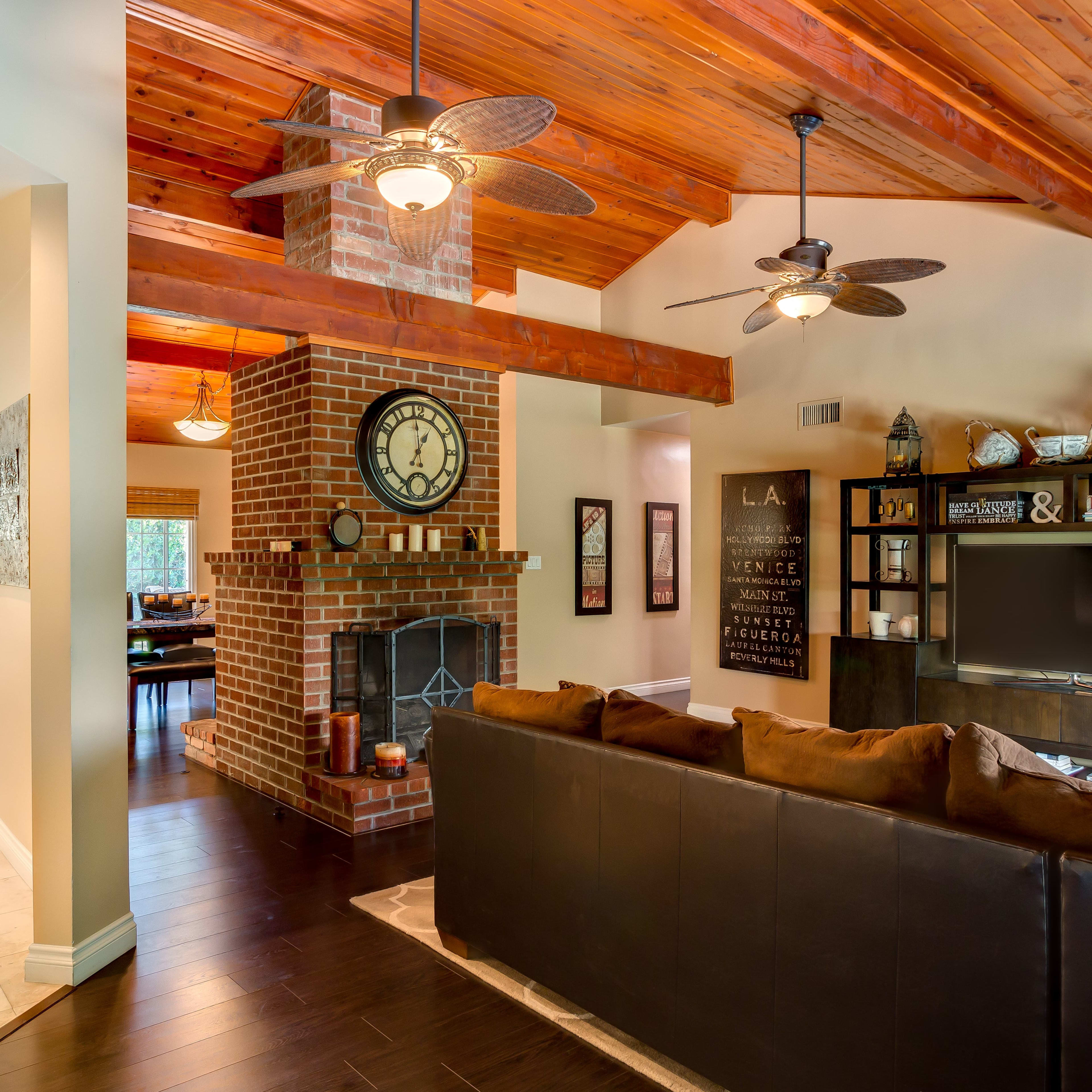 A modern living space featuring a brown sectional sofa, a brick fireplace with a clock, and wooden beams overhead, seamlessly connecting to a dining area with a kitchen.