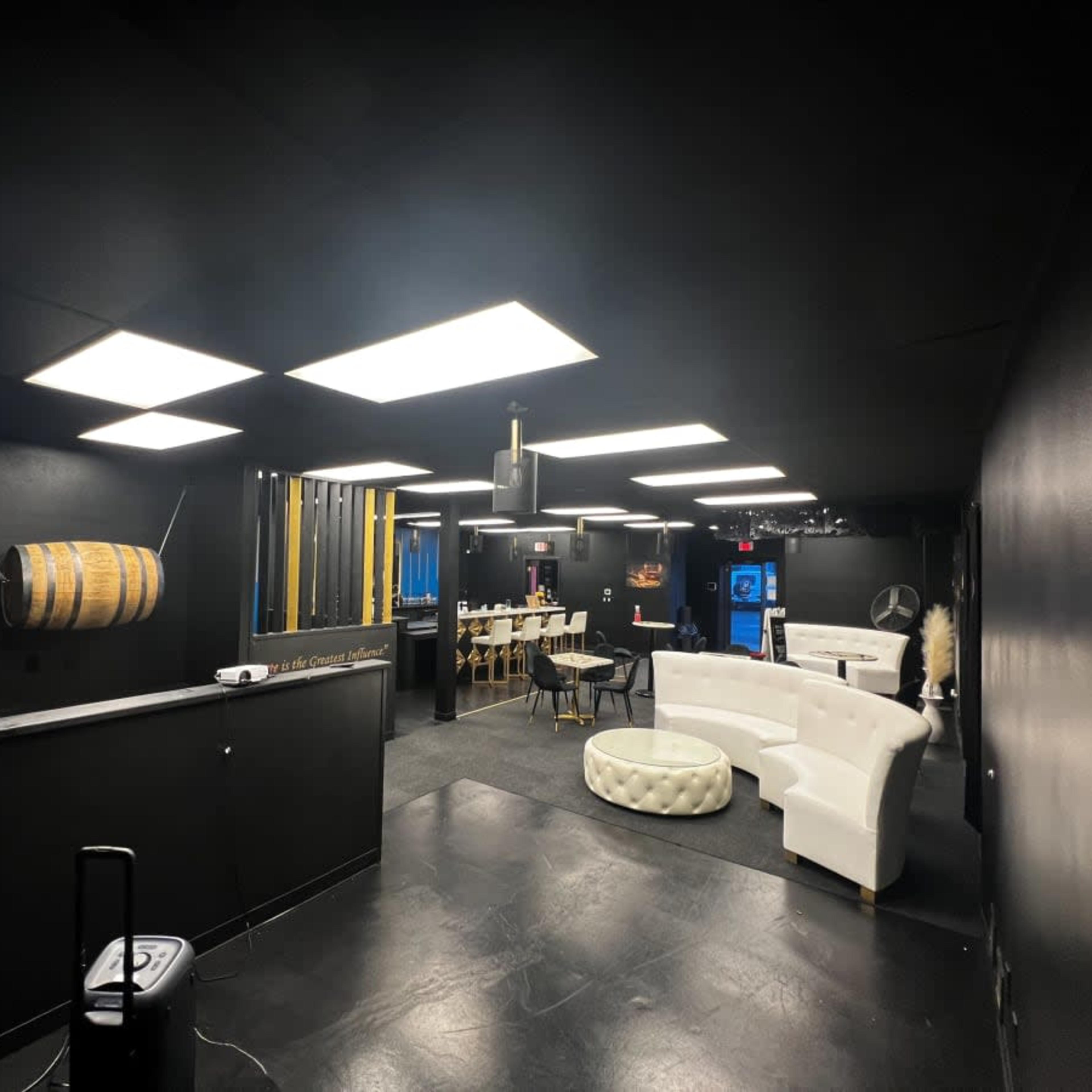 A dimly-lit lounge area with black walls, a white couch, a barrel on display, and a dining table set for several people.
