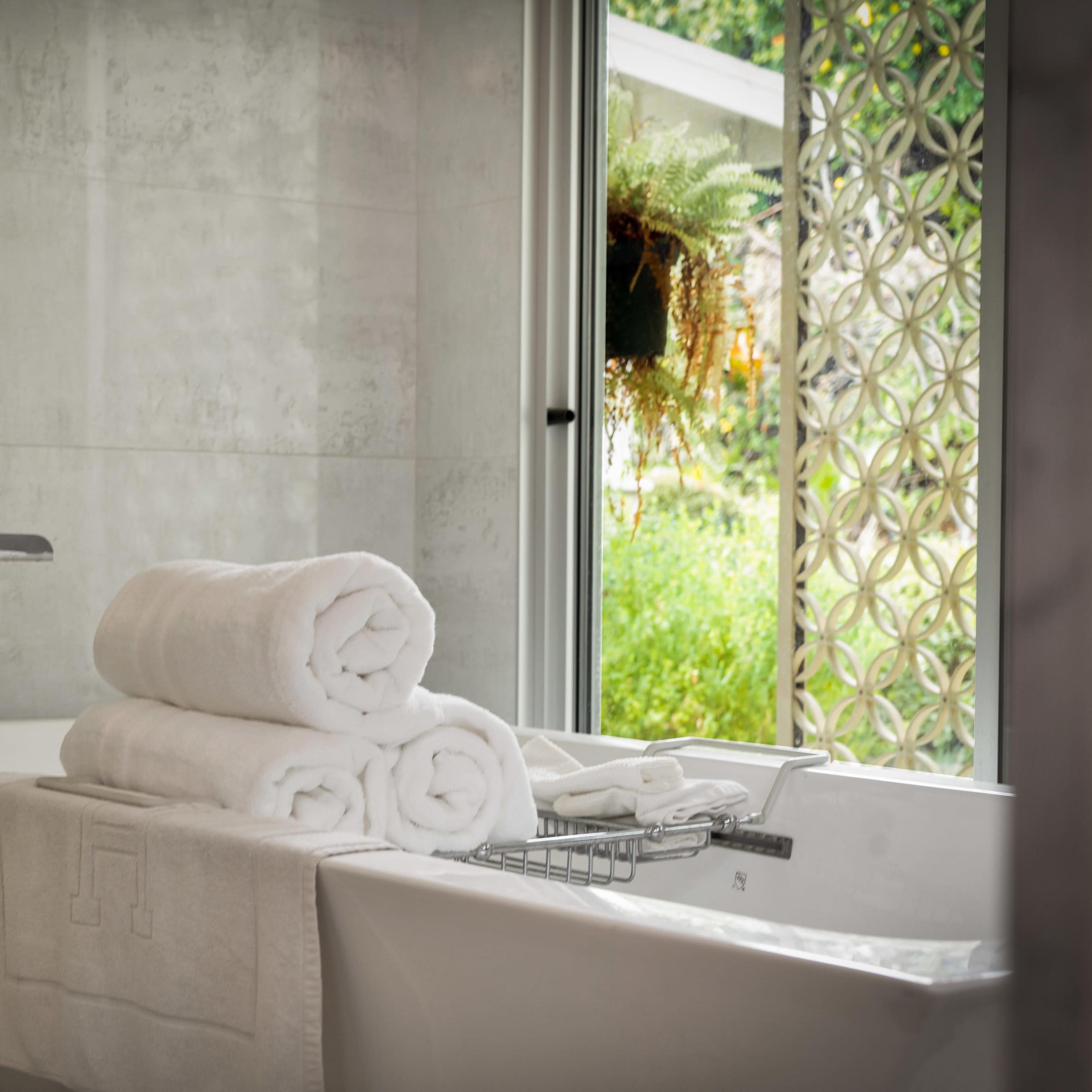 A neatly arranged stack of white towels sits on the edge of a modern bathtub, with a window displaying greenery in the background.