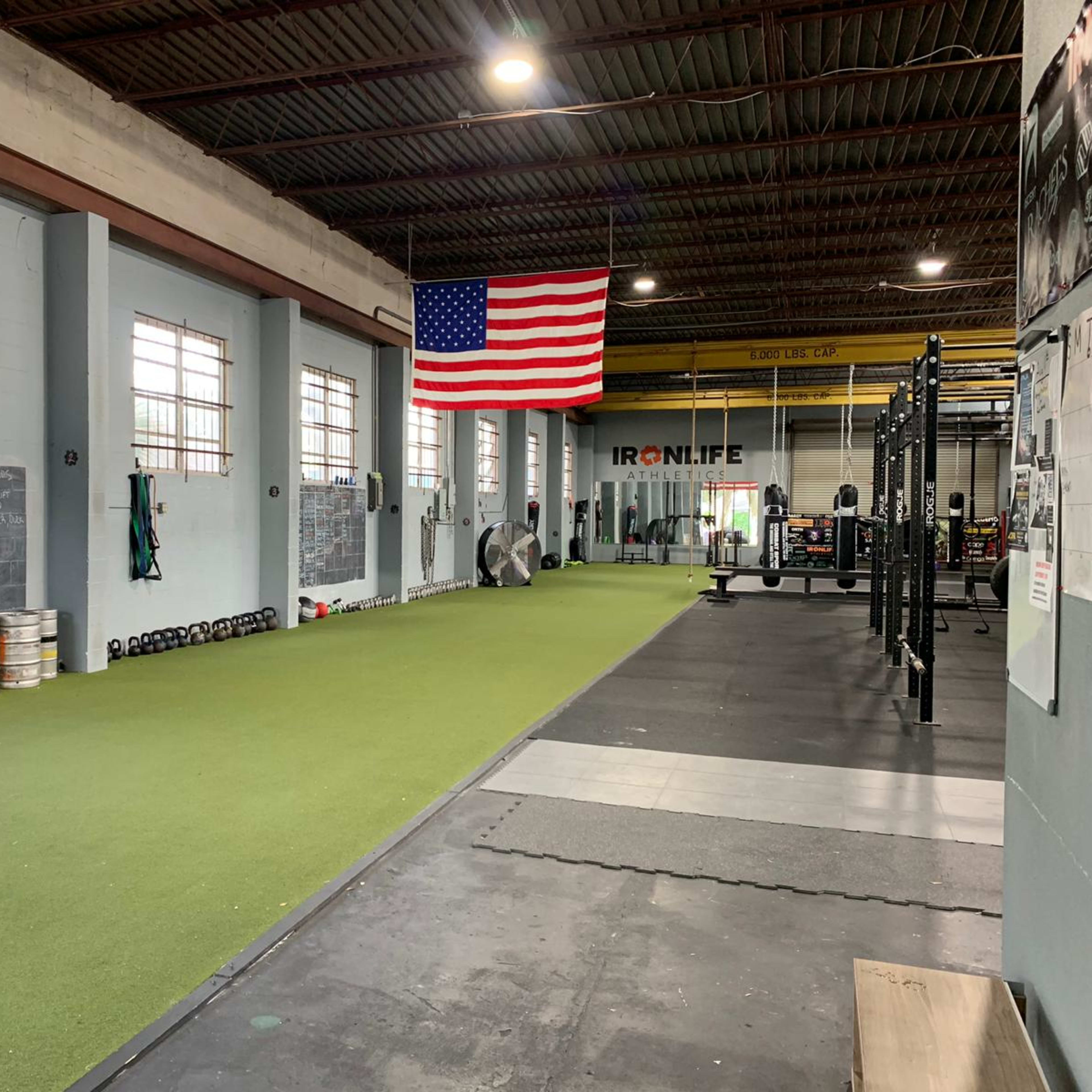 A spacious gym interior featuring green turf flooring, weightlifting equipment, and an American flag hanging on the wall.
