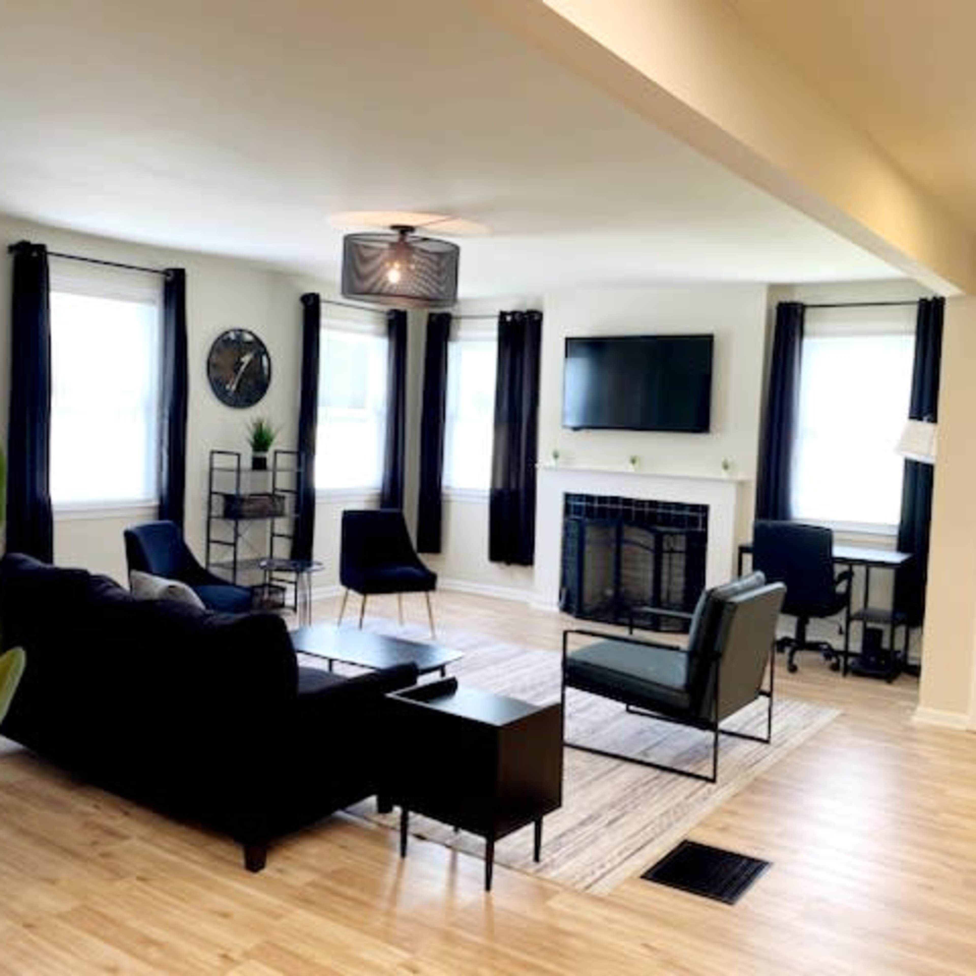The room features a modern living space with a black sofa, two chairs, a coffee table, and a desk, illuminated by natural light streaming through large windows.