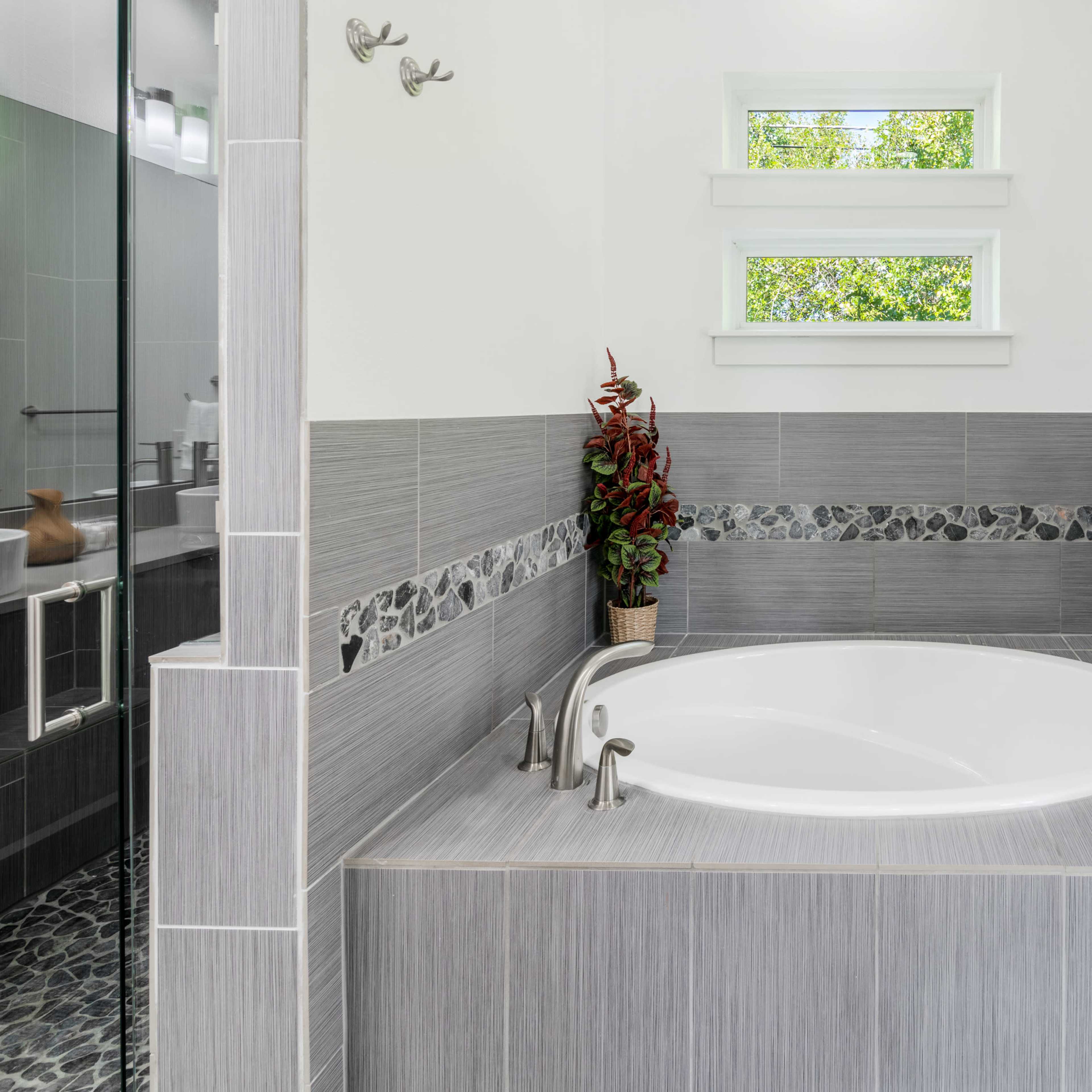 A modern bathroom featuring a round bathtub surrounded by grey tile and a decorative border, with a small plant placed on a ledge.