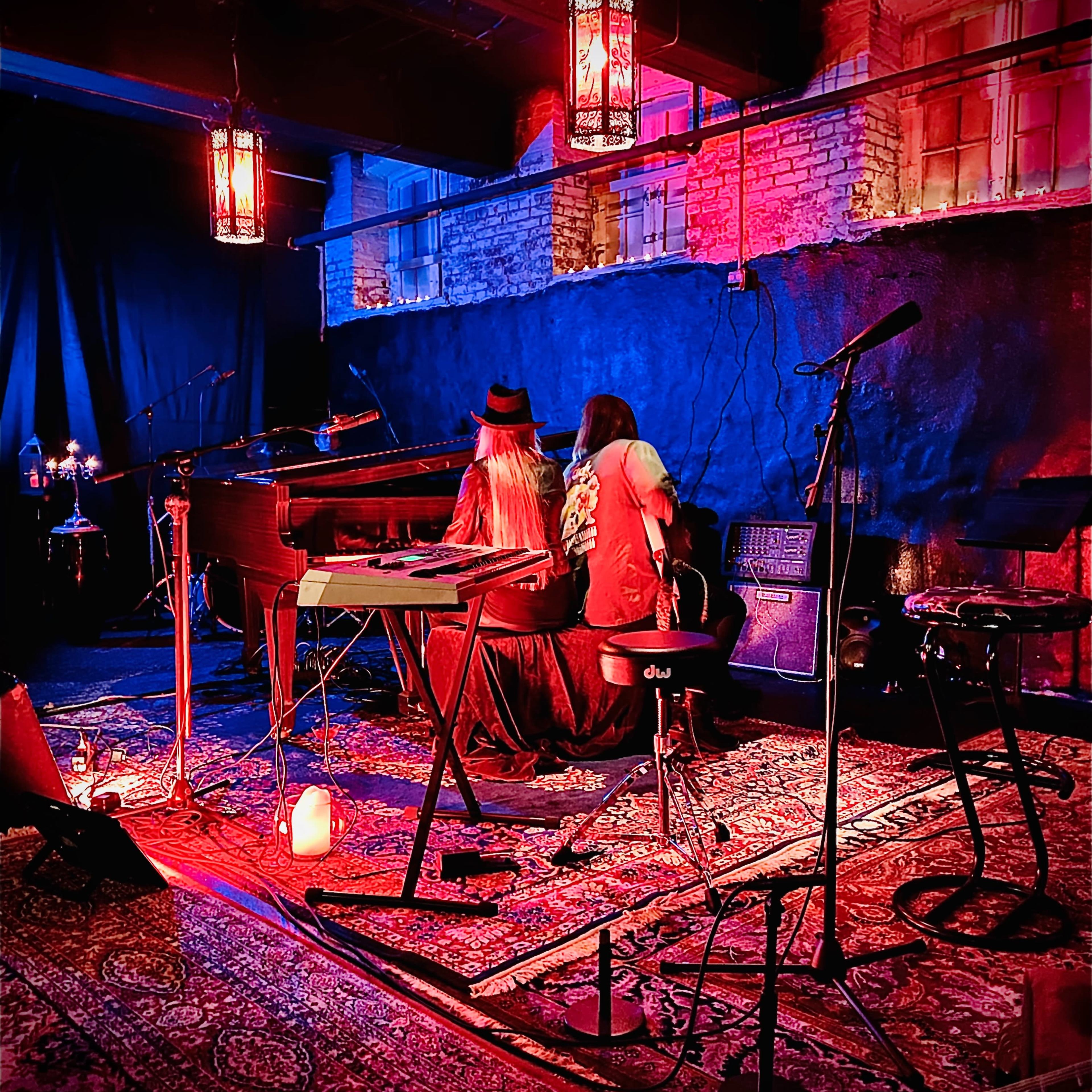 Two musicians sit at a piano and guitar in a dimly lit venue, surrounded by colorful lights and decorative lanterns.
