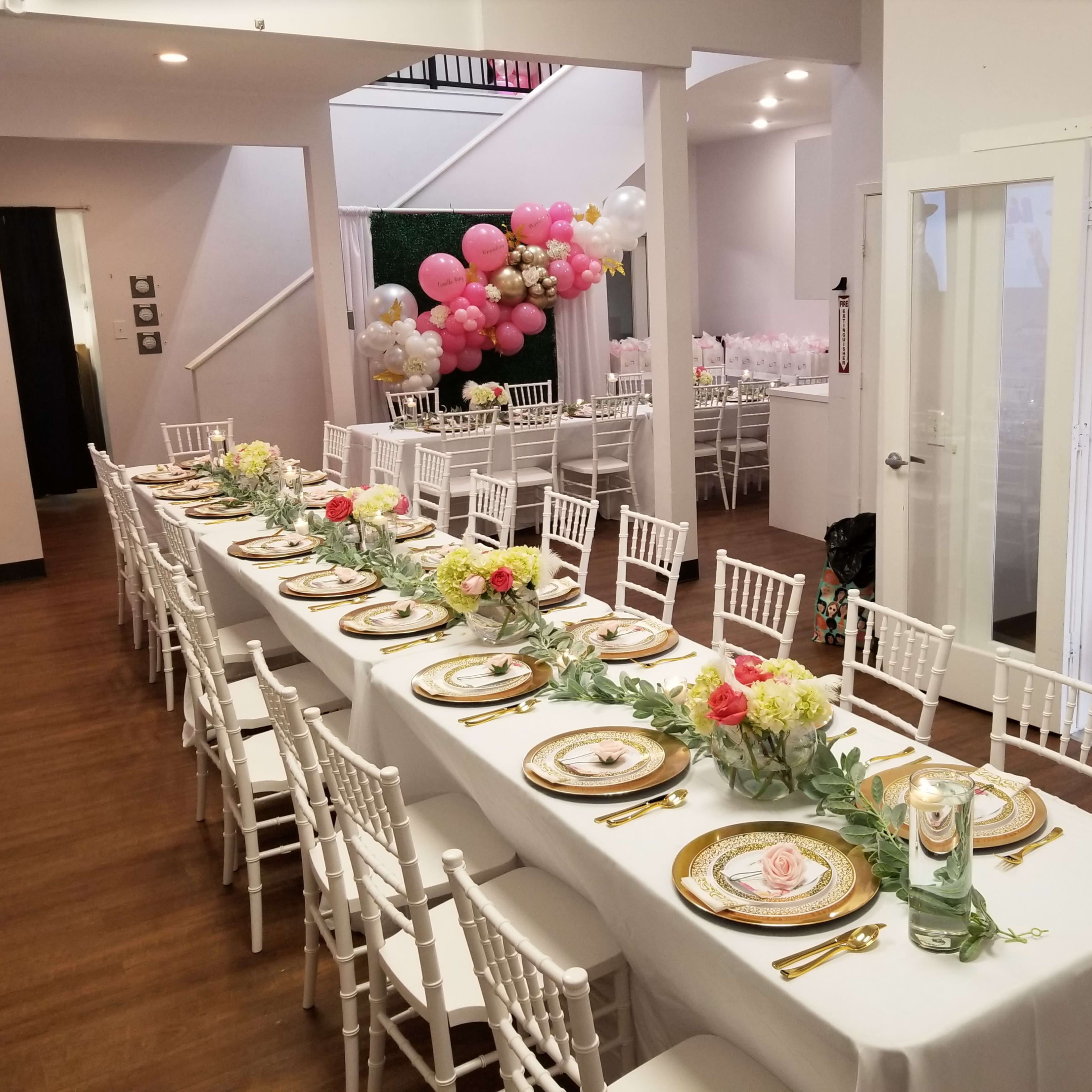 A banquet hall decorated with a long table set for an event, featuring white tablecloths, floral centerpieces, and gold dinnerware, with balloons in the background.