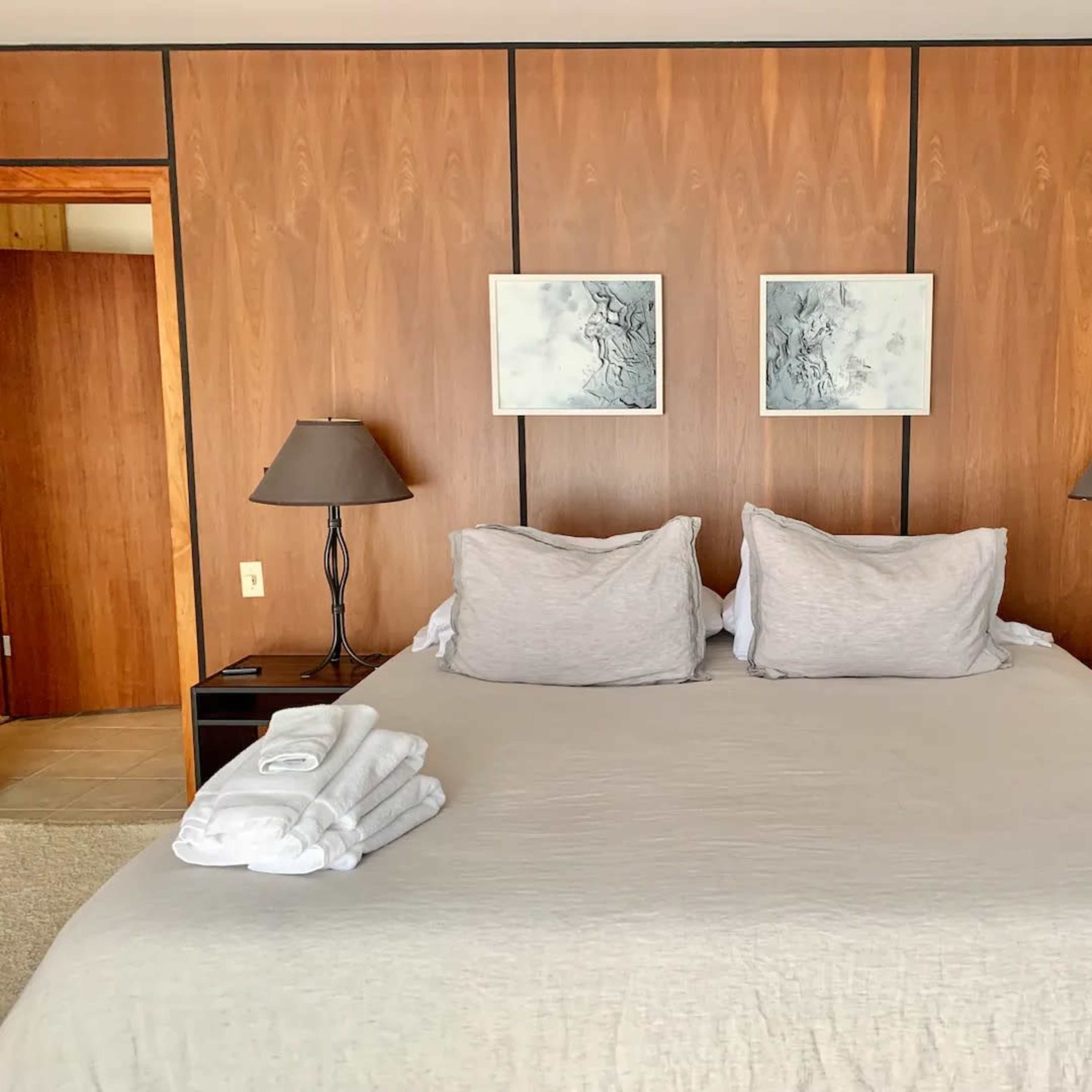 A neatly made bed with gray linens is set against a wooden-paneled wall, featuring two paintings above the headboard and bedside lamps on either side.