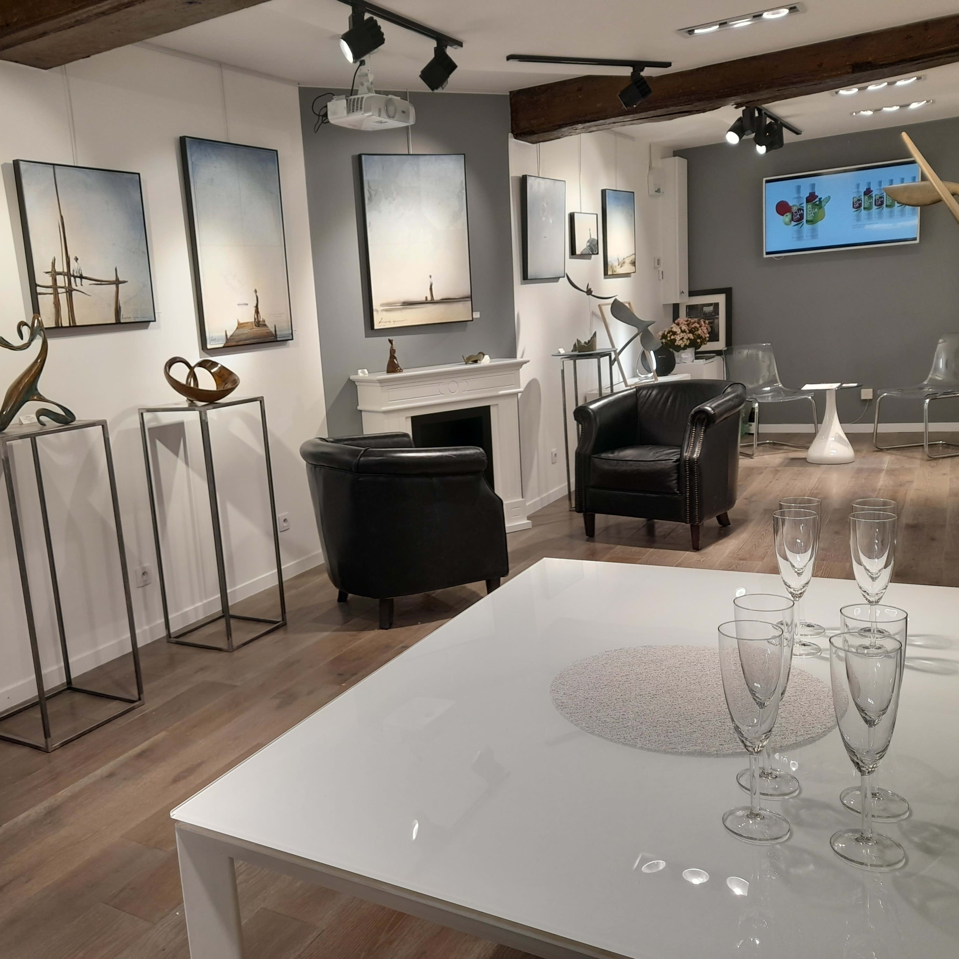 A modern art gallery featuring abstract sculptures and framed artworks on white walls, with a sleek white table set with champagne flutes in the foreground.