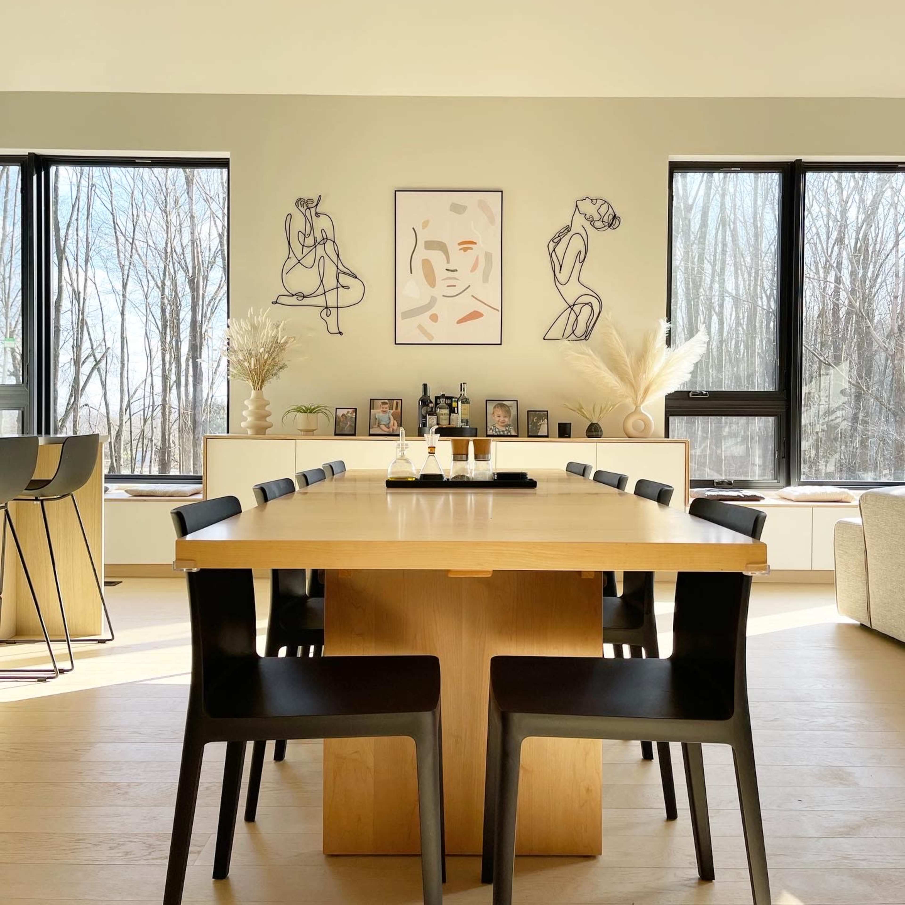 A modern dining room features a large wooden table surrounded by black chairs, with art on the walls and large windows overlooking a wooded area.