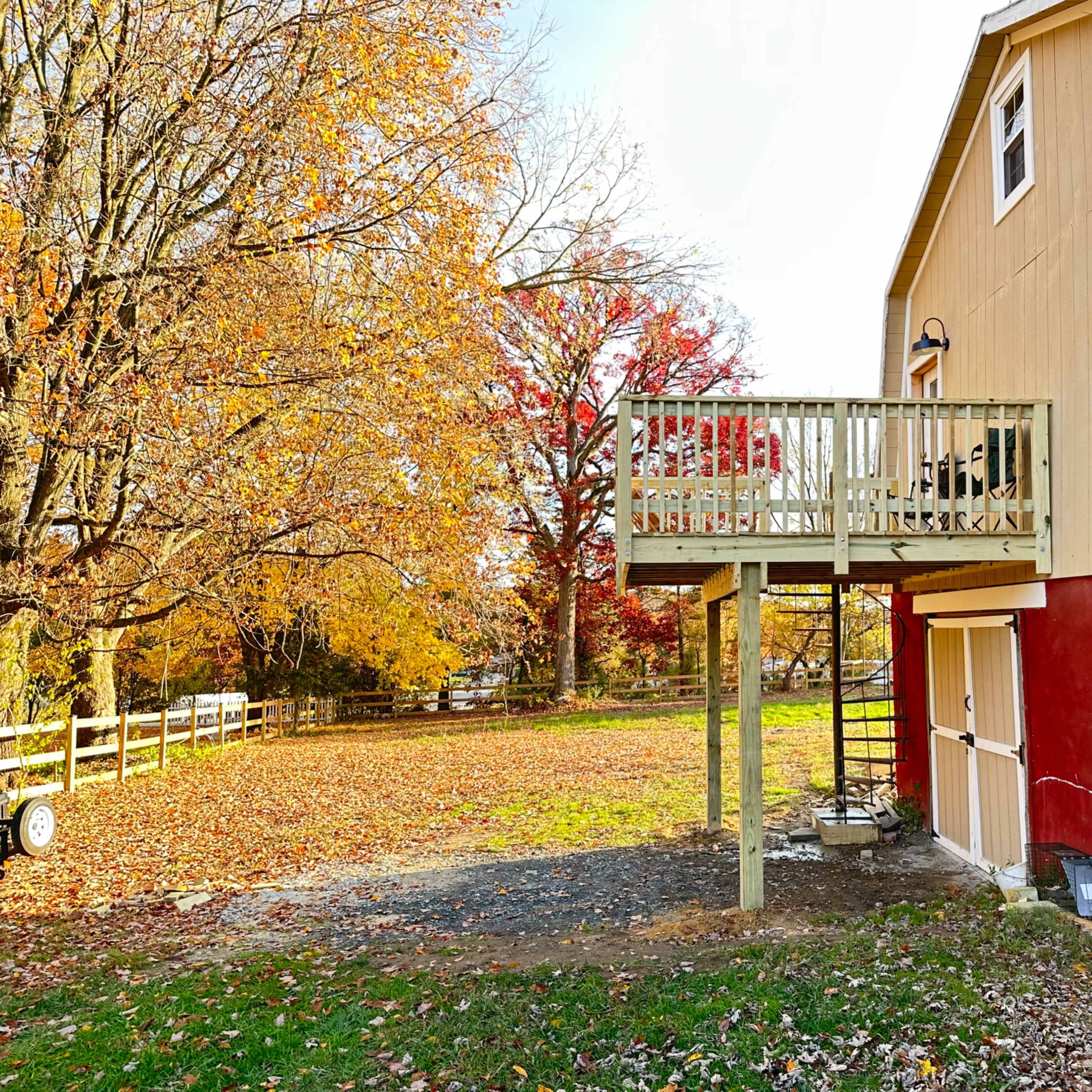 A two-story house with a wooden balcony overlooks a yard covered in autumn leaves, surrounded by trees.