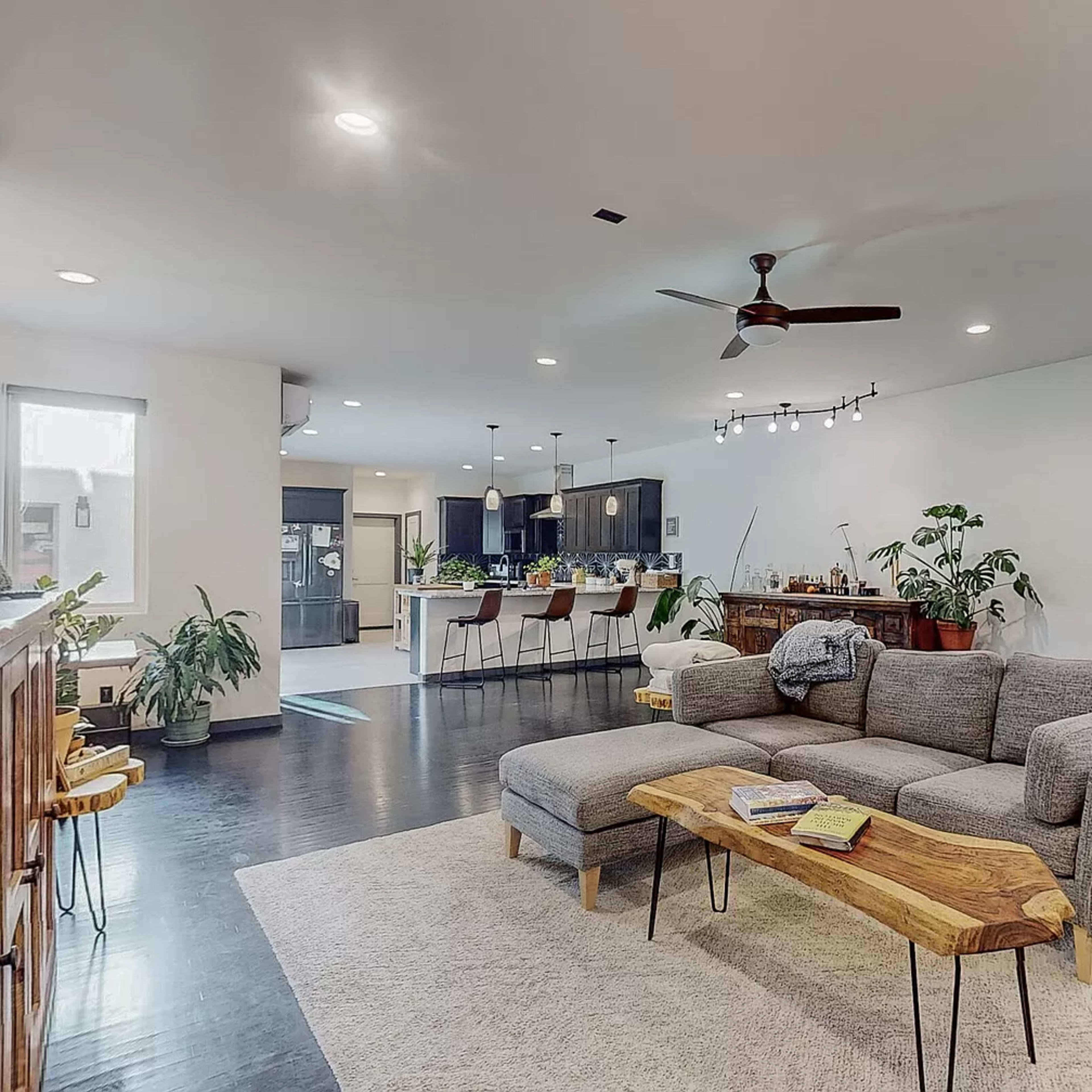 An open-concept living area featuring a grey sectional sofa, a natural wood coffee table, and a kitchen with bar stools in the background.