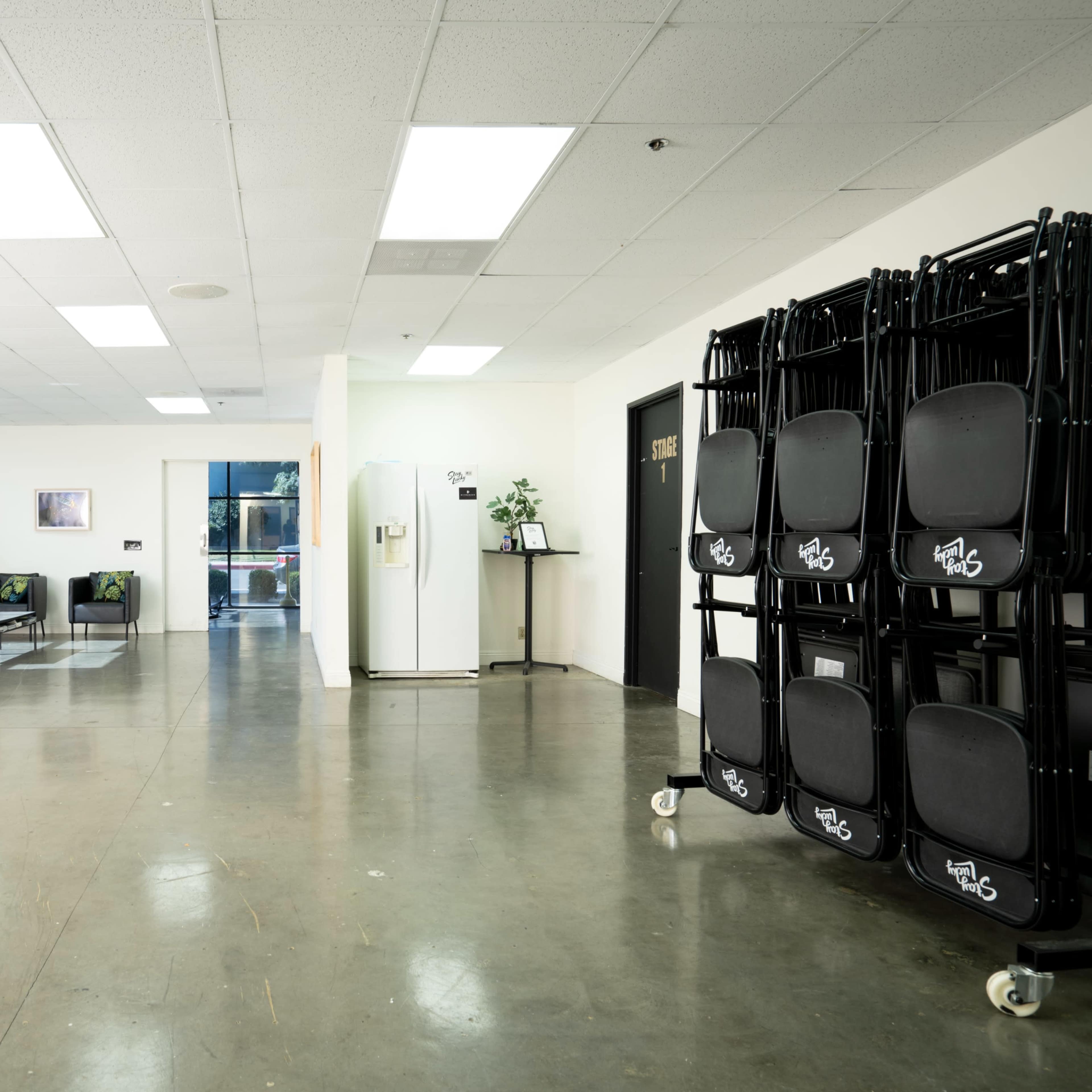 An open room with concrete flooring, a lounge area featuring a black sofa and plants, and a stack of folding chairs against a wall.