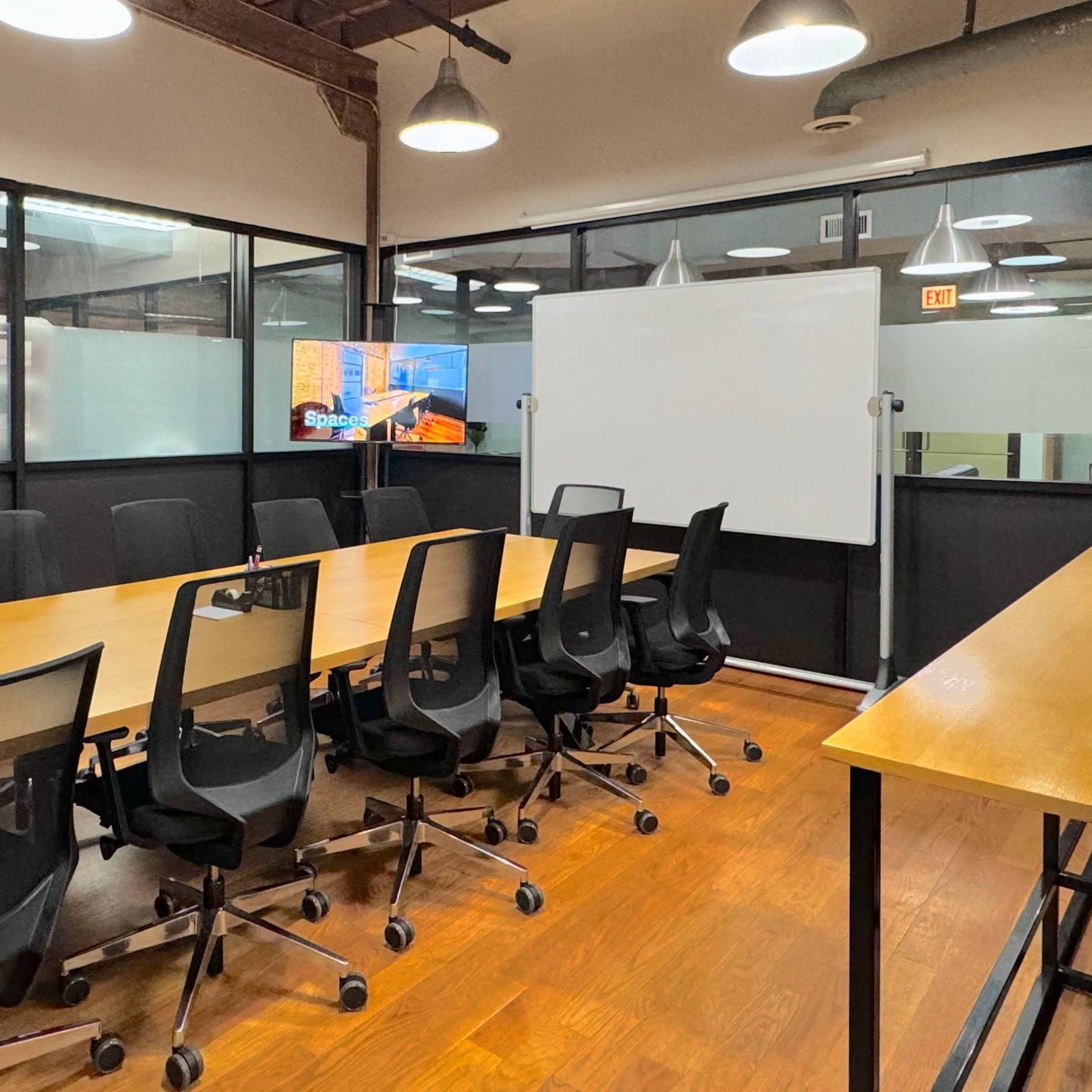 A modern conference room features a long wooden table surrounded by black swivel chairs, with a large screen mounted on the wall and a whiteboard nearby.