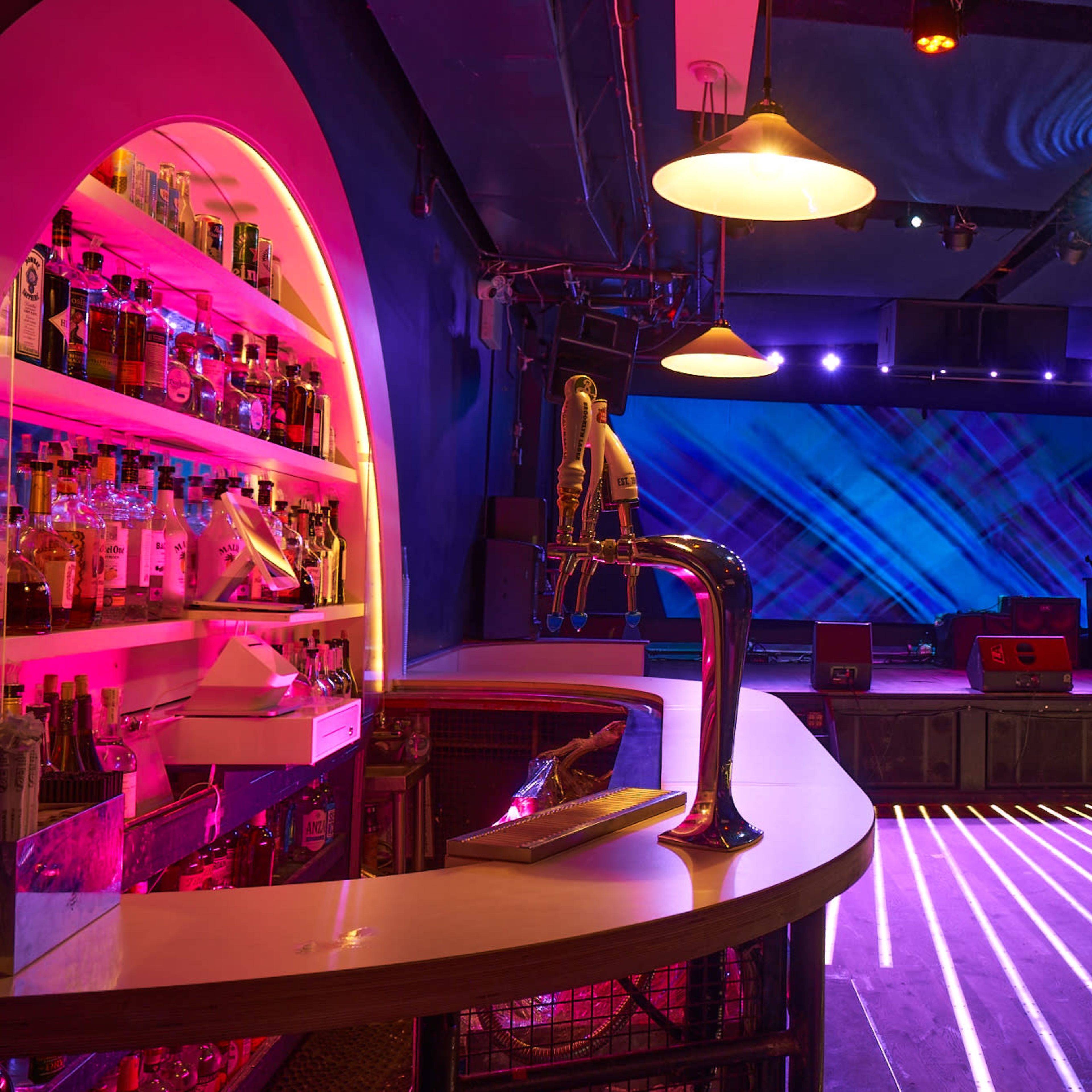 A bar area in a dimly lit venue with a colorful backlit display, shelves of liquor, and stage setup with lighting effects.