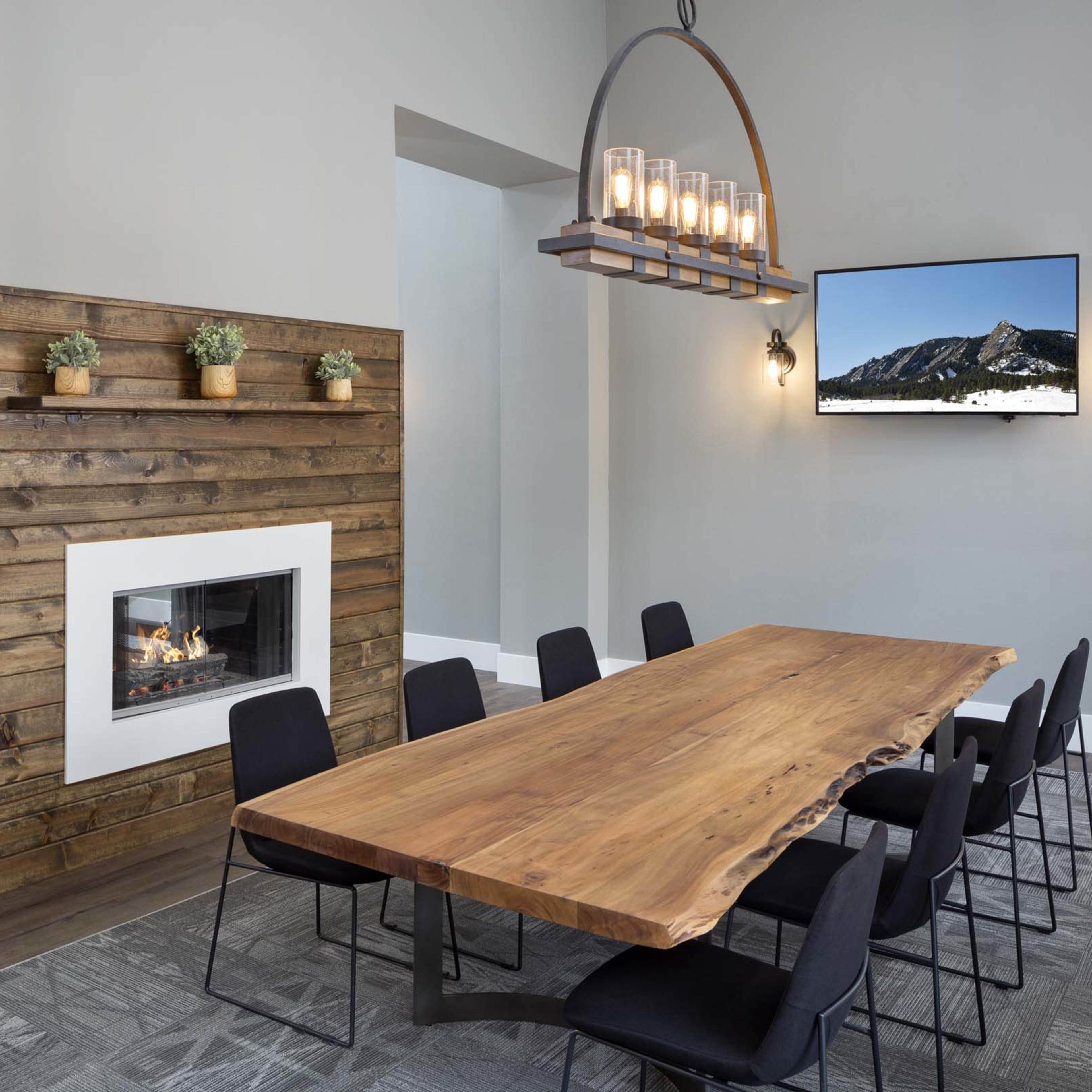 A large wooden dining table surrounded by black chairs is set in a modern room featuring a fireplace and a mounted television on gray walls.