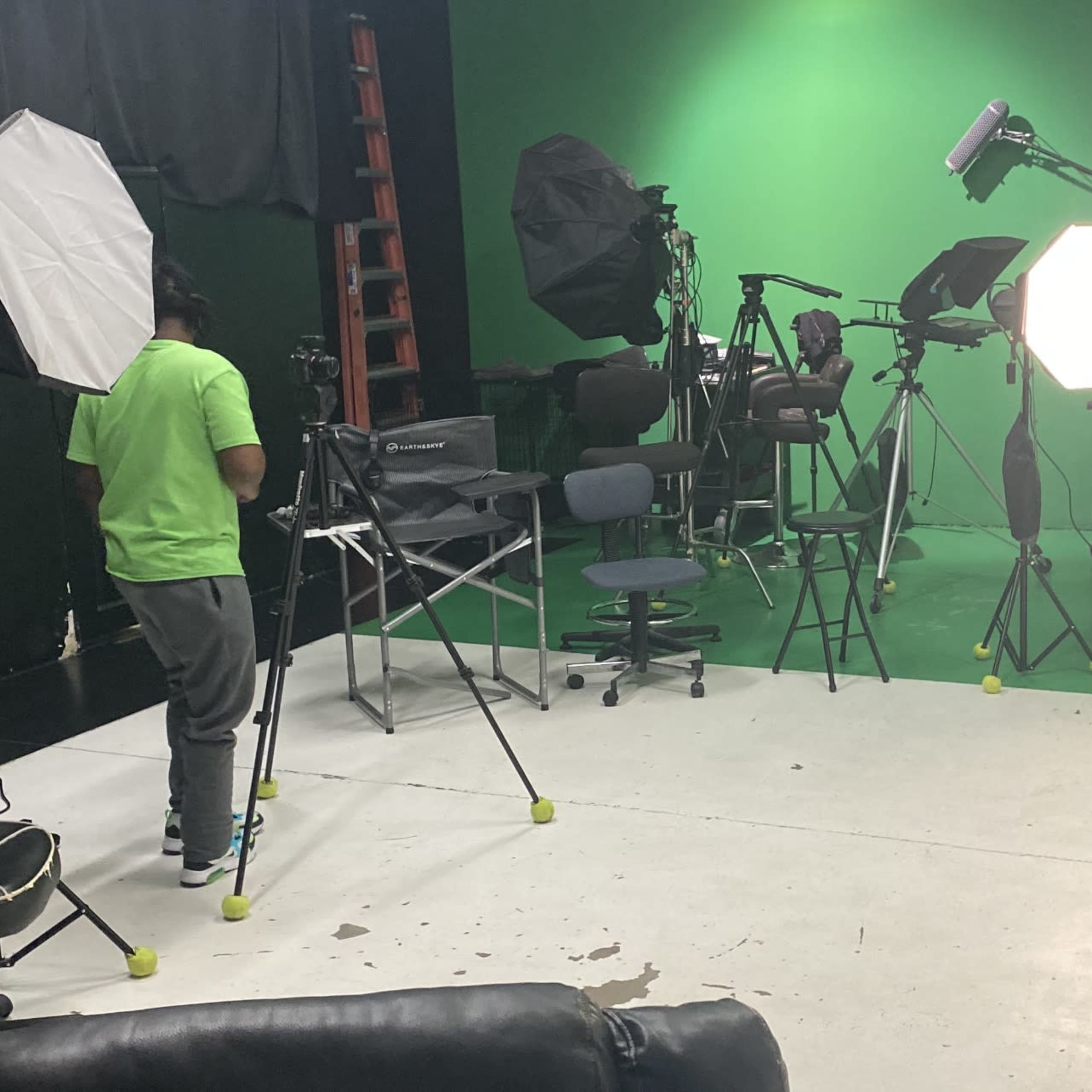 A person stands in a studio with a green backdrop, surrounded by various photography equipment, including lights and tripods.