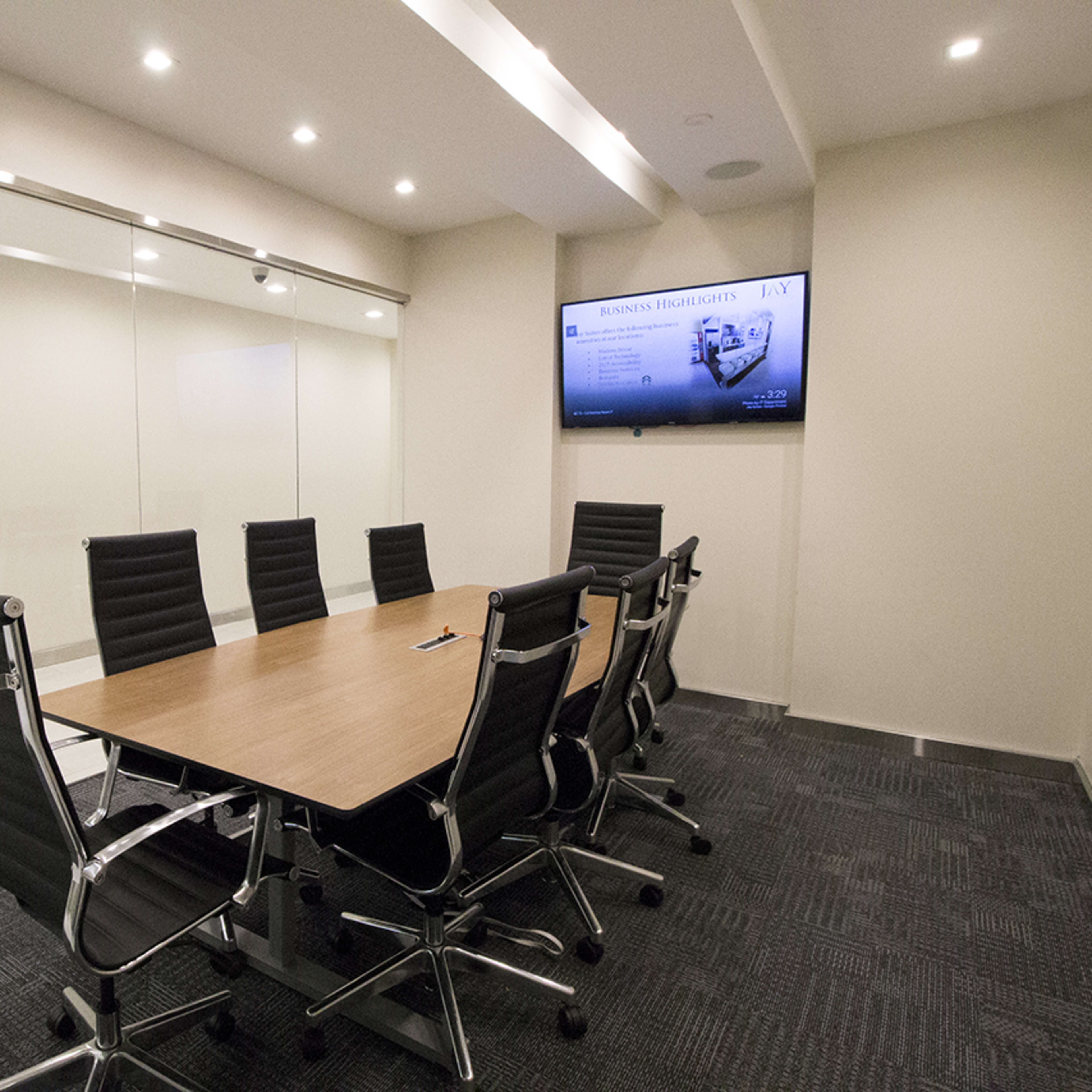 A conference room features a large wooden table surrounded by black leather office chairs, with a wall-mounted screen and glass walls.
