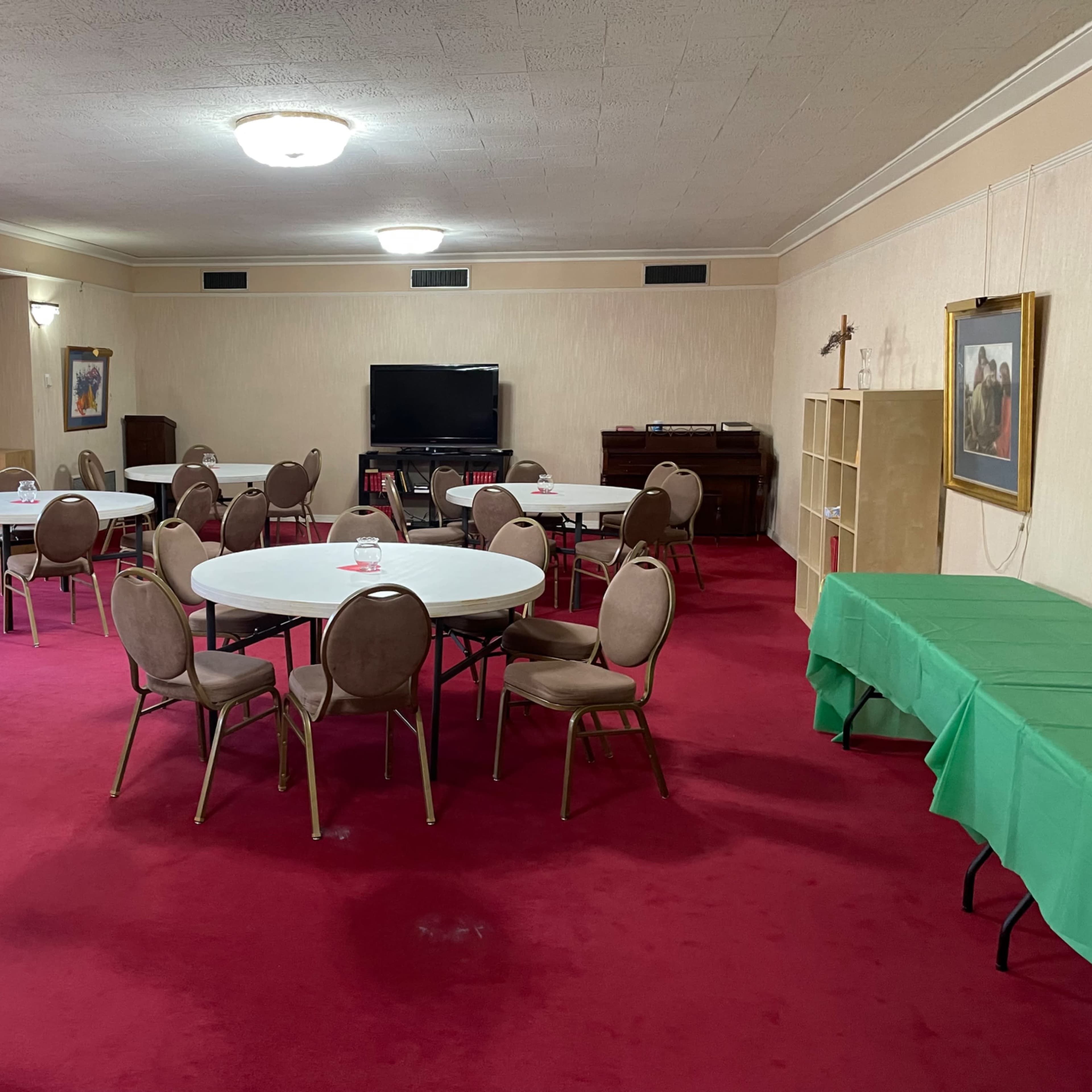 A large, carpeted room features several round tables with chairs, a television, and a piano along one wall.