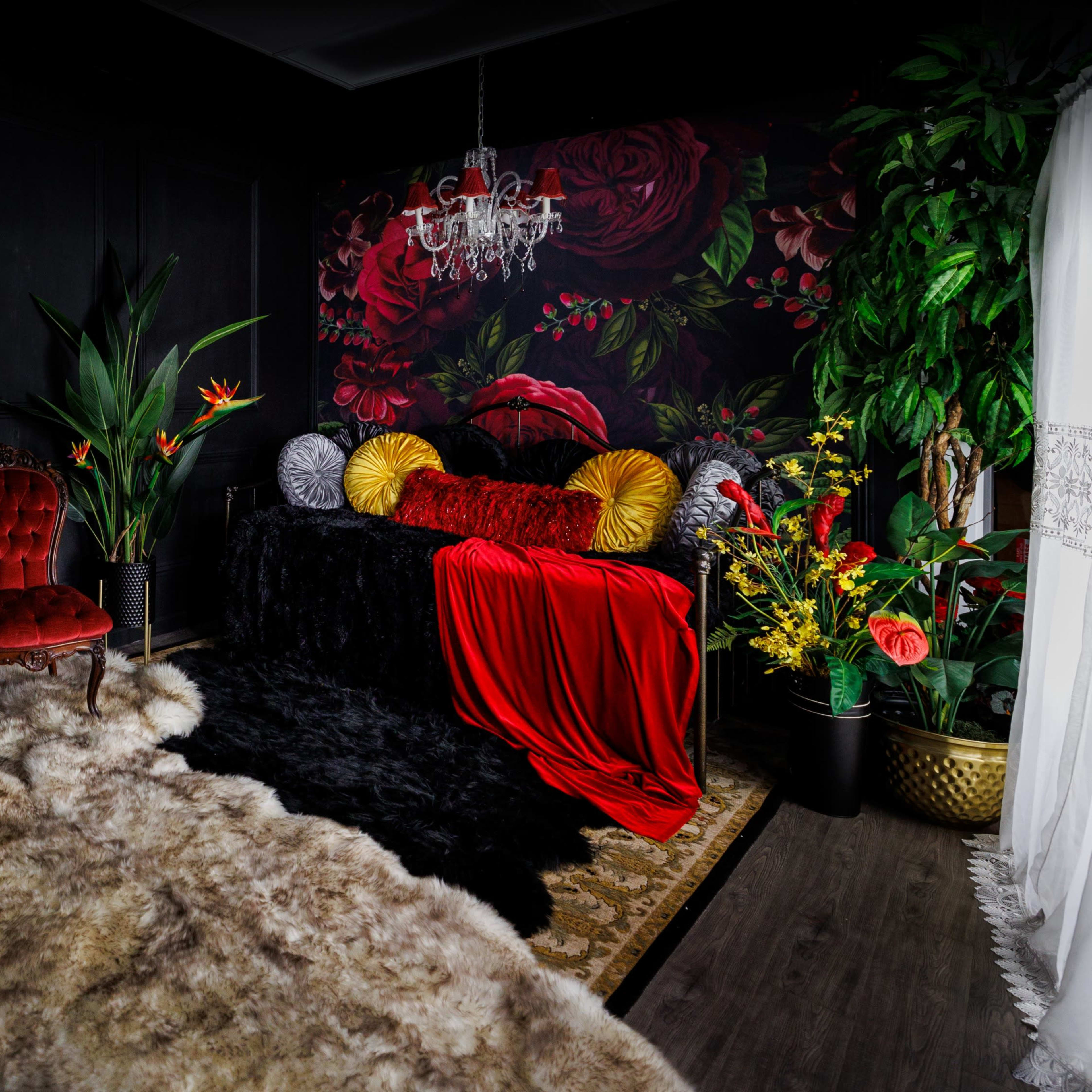 An elegantly styled room featuring a black wall adorned with floral wallpaper, a plush fur rug, a vintage chair, a decorative chandelier, and vibrant floral arrangements.