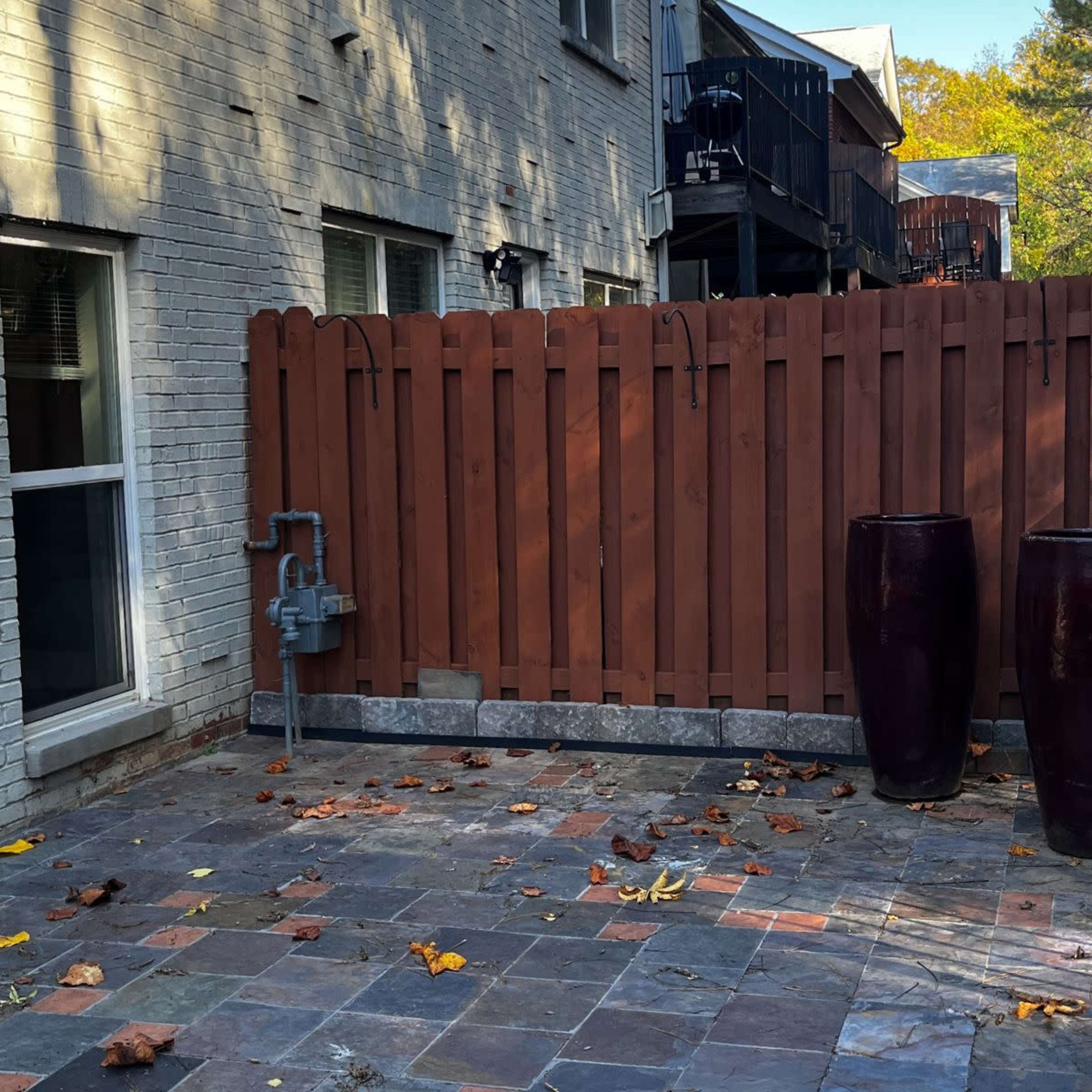 A fenced outdoor area with two dark planters and a stone pavement, surrounded by a residential building and trees in the background.