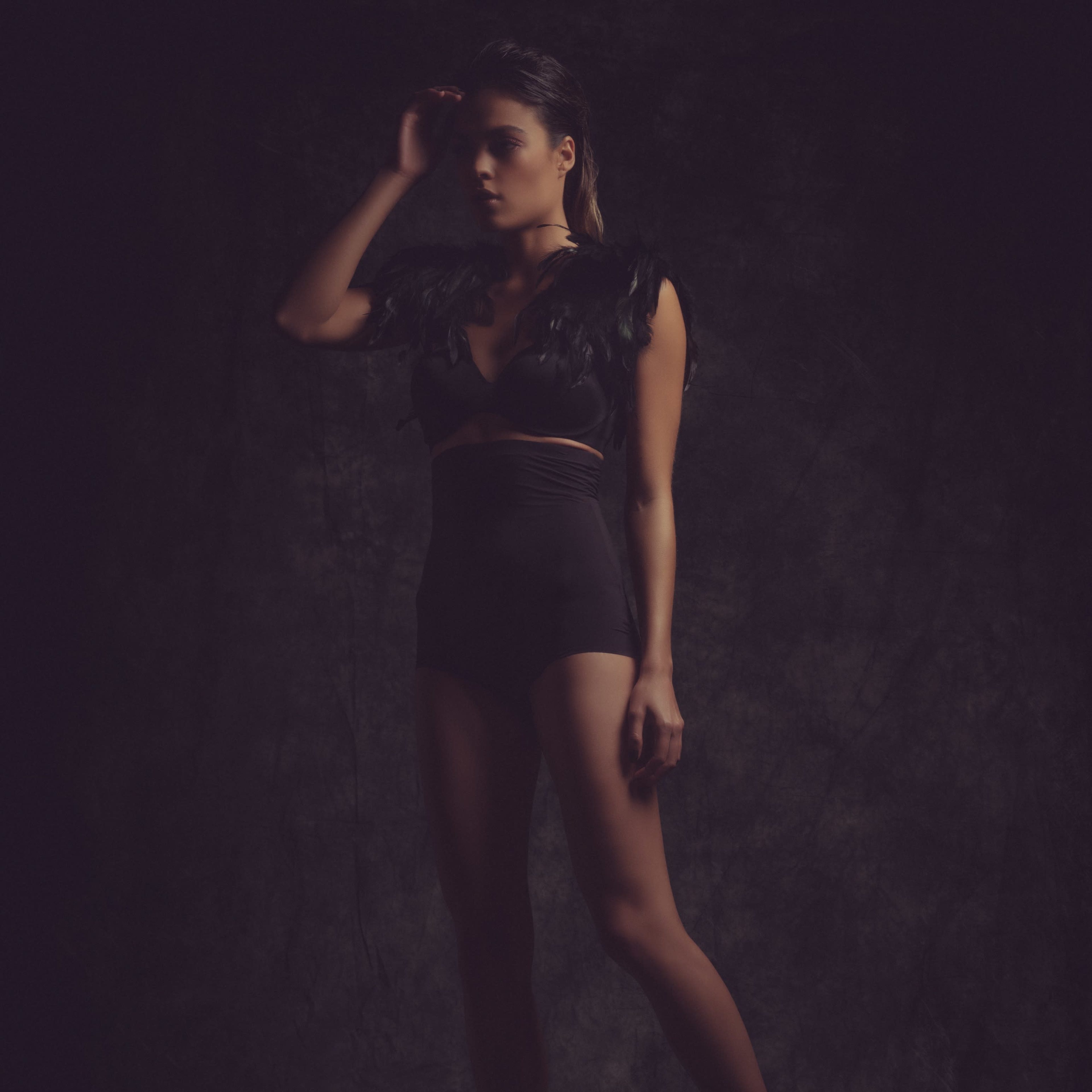 A model poses in a dark setting, wearing a black outfit with feathered accents and high heels.