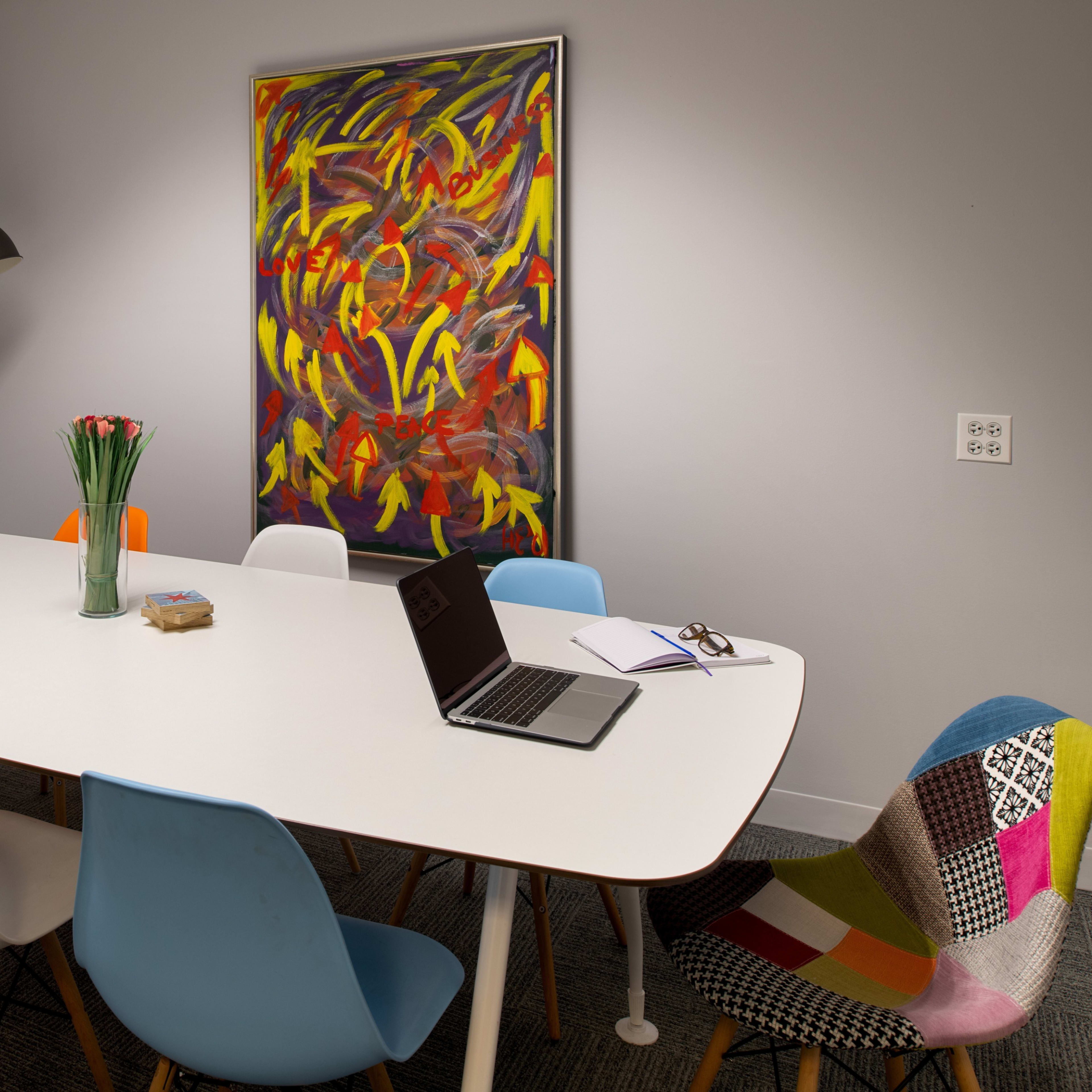 A modern conference room features a long white table surrounded by multicolored chairs, with a laptop and notebook on the table, and a large abstract painting on the wall.