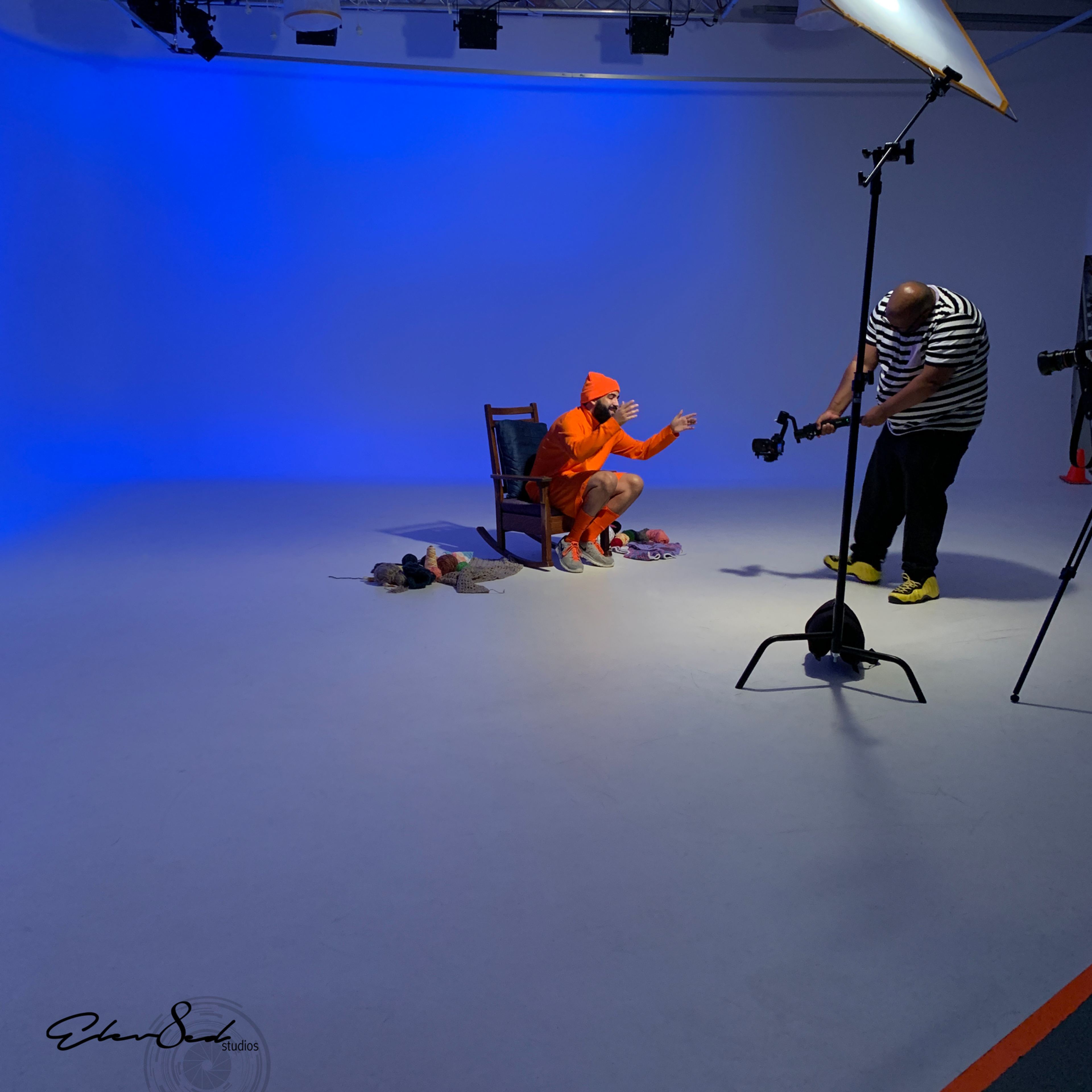 A person in an orange outfit sits on a chair while another individual adjusts a camera in a photography studio with blue lighting.