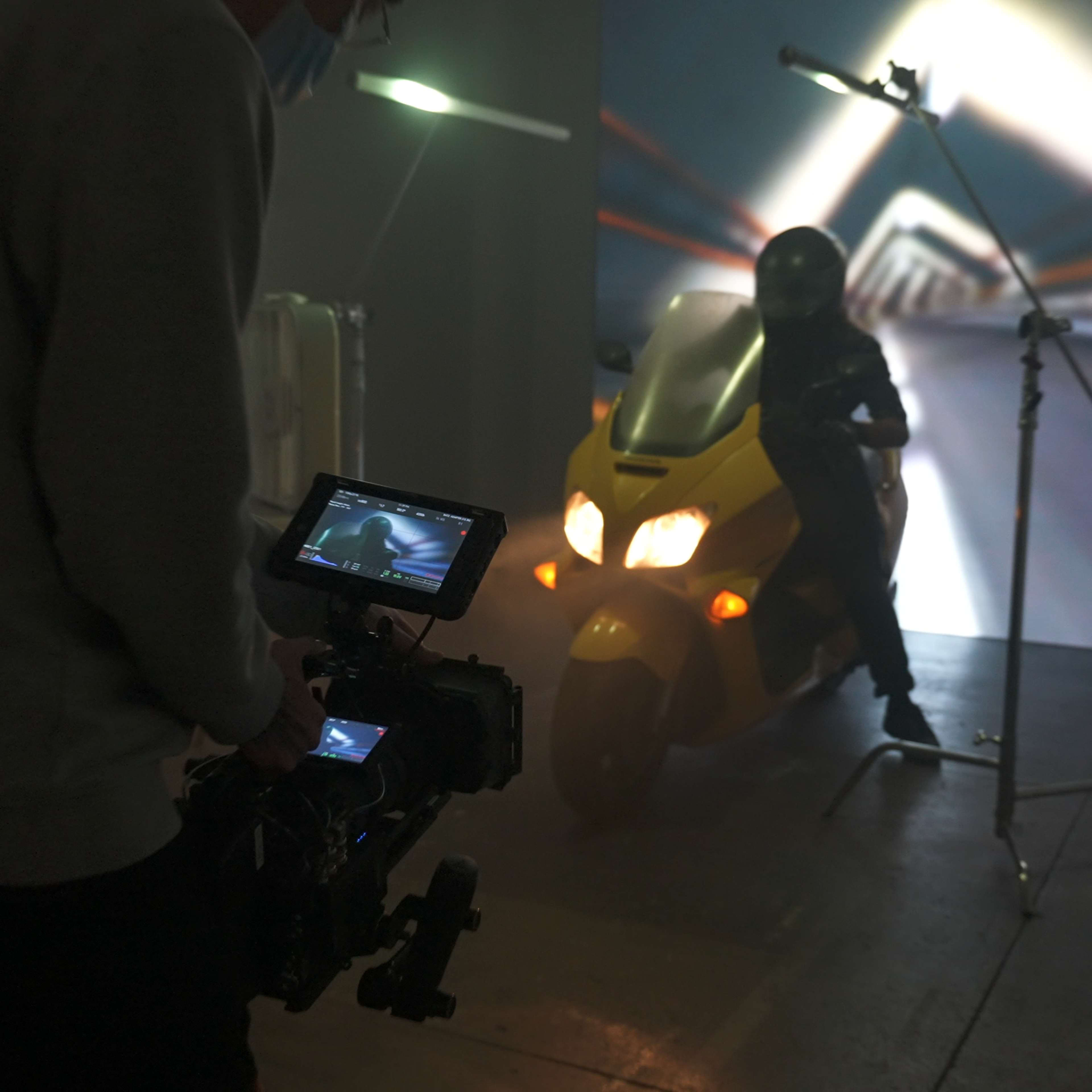 A cameraman operates a camera while filming a person on a yellow motorcycle in a studio with a projected background.