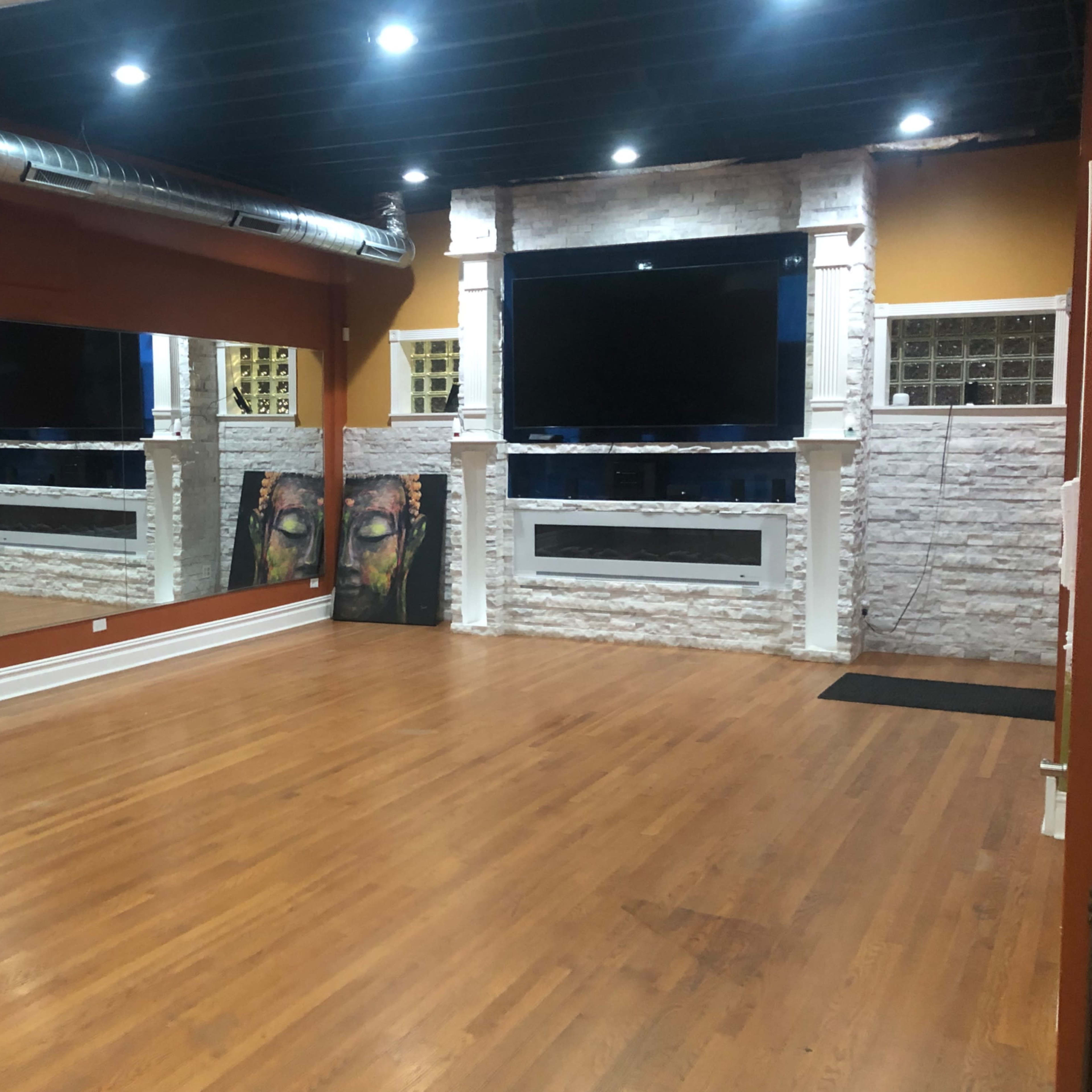 An indoor studio space with wooden floors, a large mirror on one wall, and a prominent flat-screen TV mounted above a white stone feature wall.